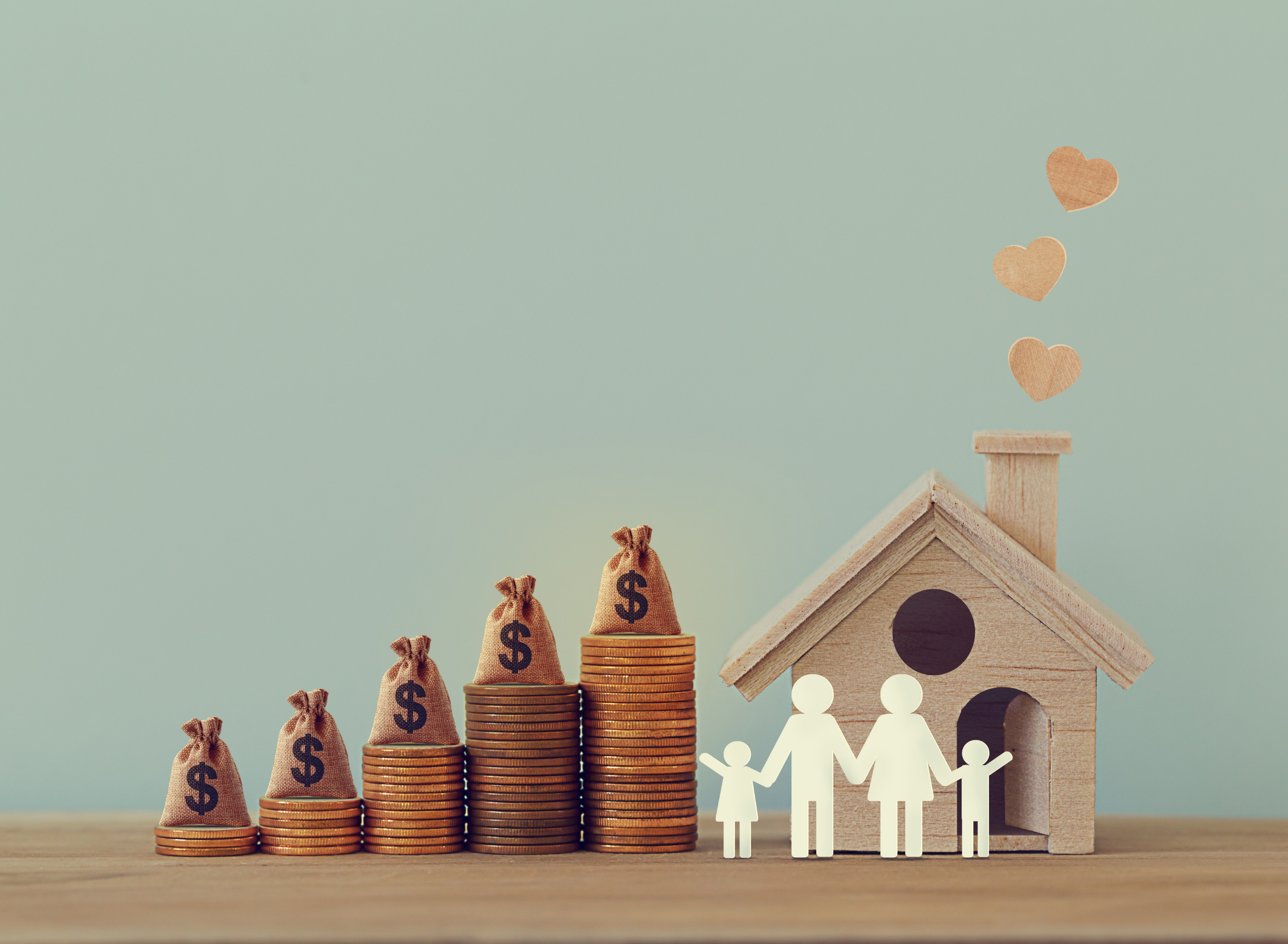 Stacks of coins increasing in height with small money bags on top, wooden family figures holding hands in front of a wooden house, with paper hearts floating above.