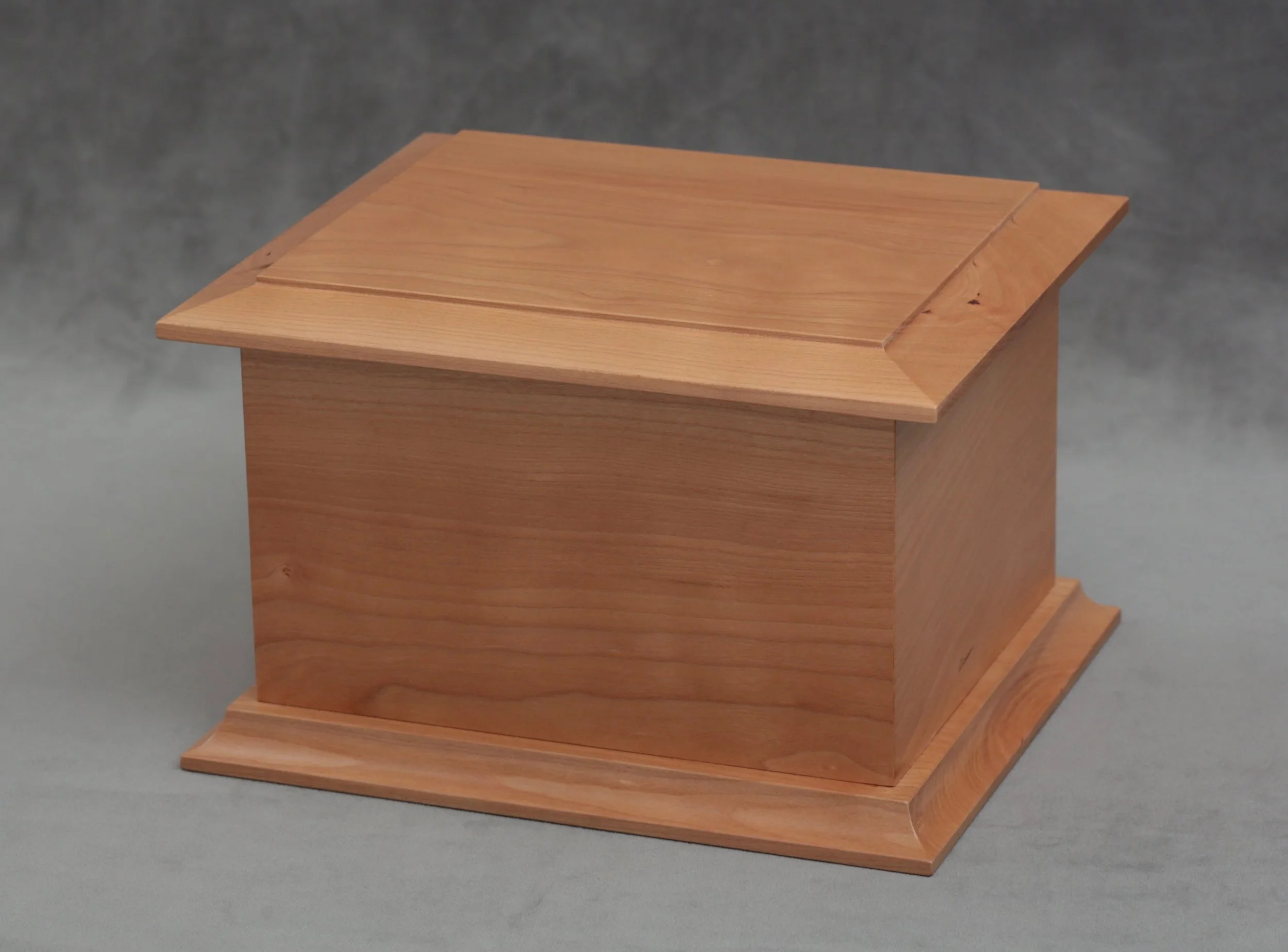 A small wooden box with a sloped lid, made from light-colored wood with visible grain, resting on a gray surface against a gray background.