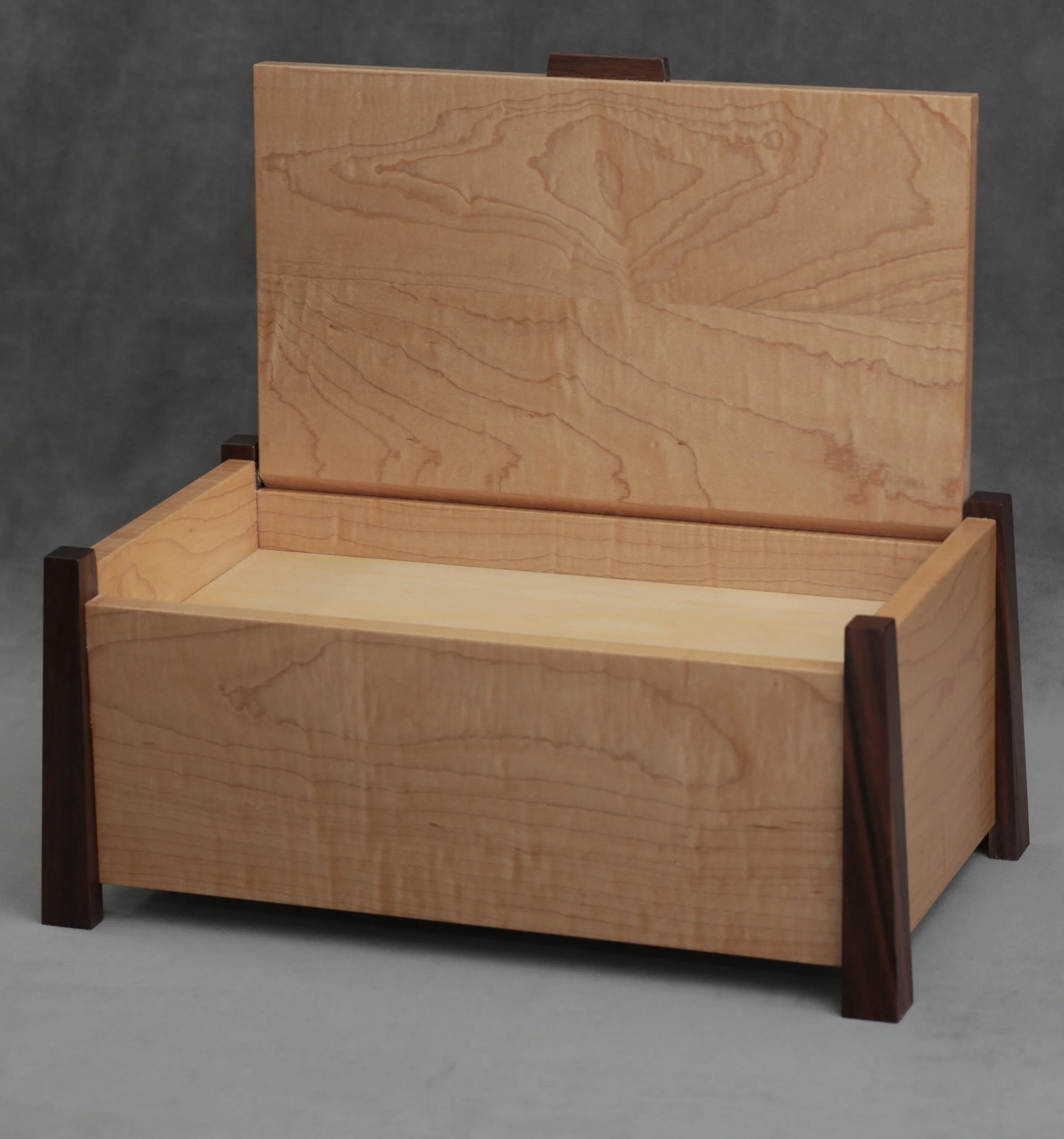 A wooden box with a hinged lid, made from light-colored and dark-colored wood, on a gray surface with a dark background.