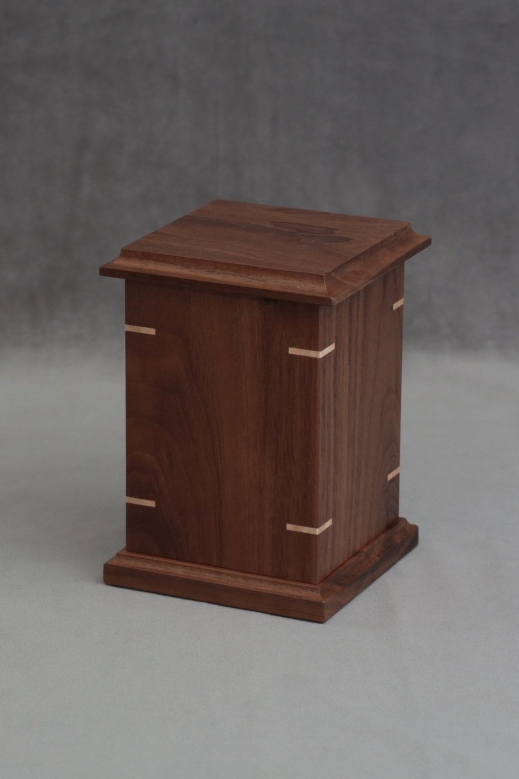 A small, rectangular wooden box with a sloped top and decorative light wood inlays on the sides, sitting on a gray surface with a darker gray background.