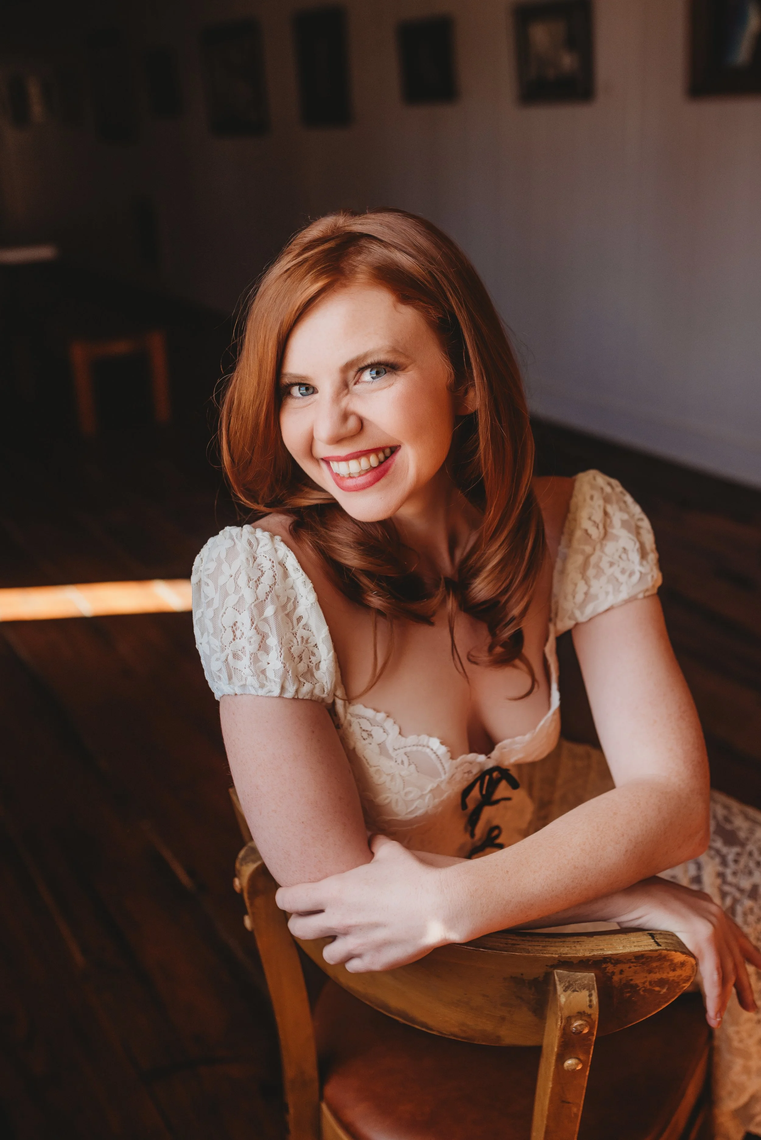 A woman with red hair, blue eyes, and a bright smile, wearing a lace cream-colored dress, sitting on a wooden chair in a warmly lit room with framed photographs on the wall.