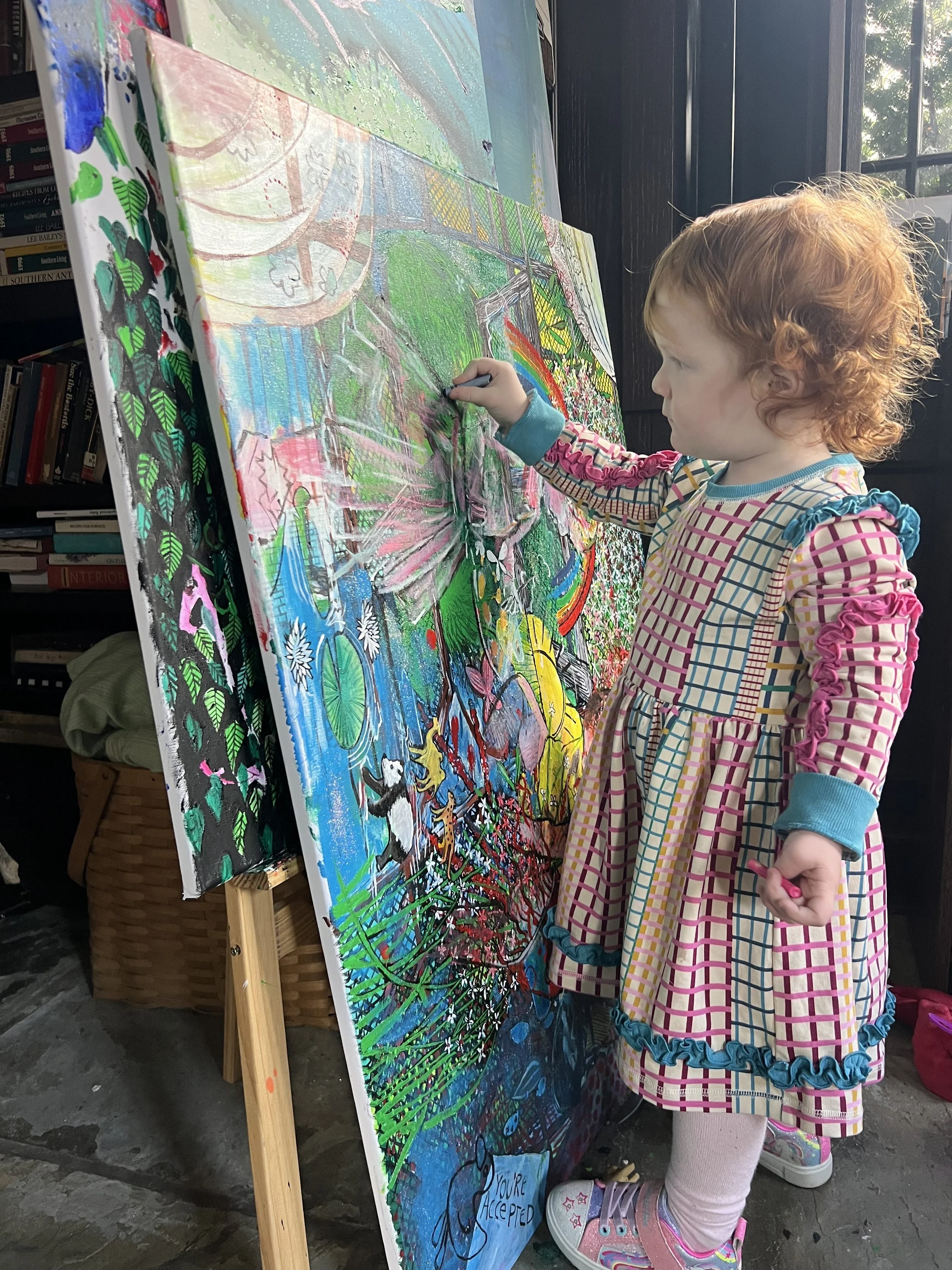A young girl with curly red hair wearing a colorful checkered dress, pink tights, and striped sneakers, is painting on a large canvas with bright colors. She is using a small brush and stands in an indoor space near a window, with books and a basket visible in the background.