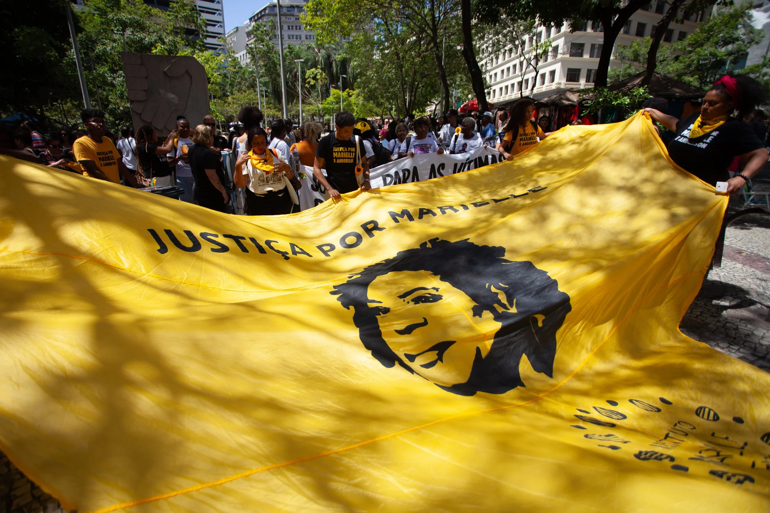 Protest march with a large yellow banner displaying a black and white portrait of Marielle Franco and the text 'Justiça por Marielle' amidst a crowd of people in Rio de Janeiro surrounded by trees and tall buildings.