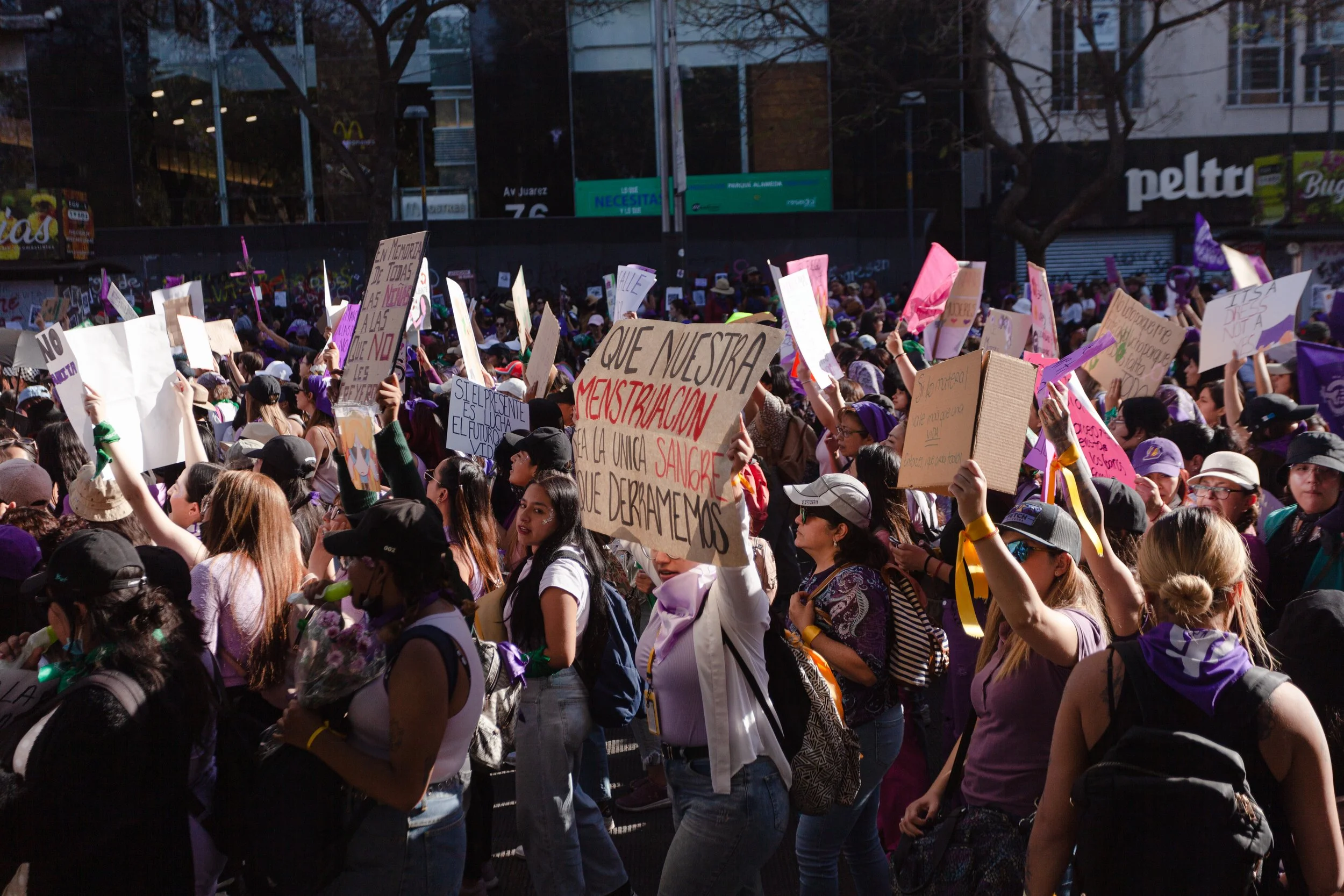 A large crowd of women protesting, holding up signs and banners, with some wearing purple and green bandanas, in an outdoor urban setting during daytime.
