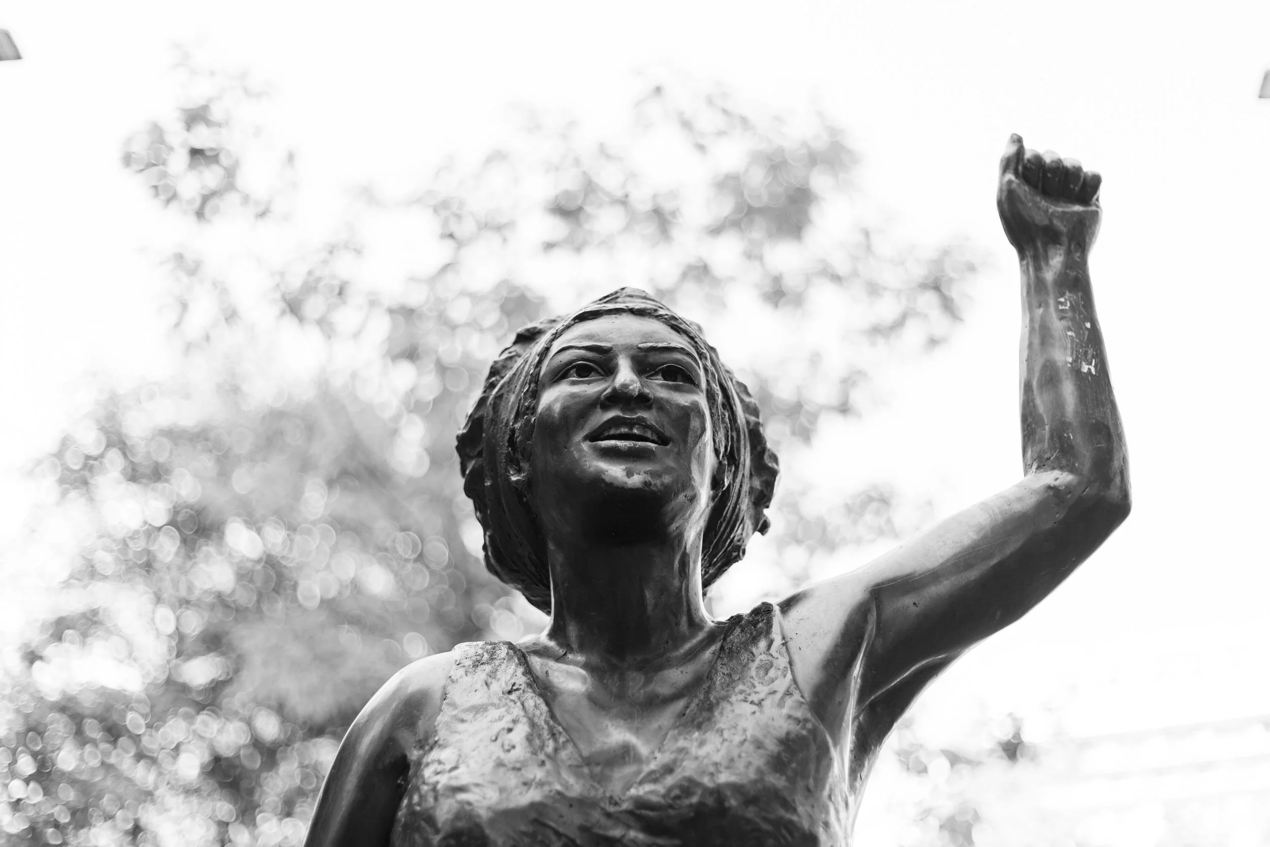 Black and white photo of a statue of Marielle Franco, holding her right arm up in a fist, outdoors with trees and sky in the background.