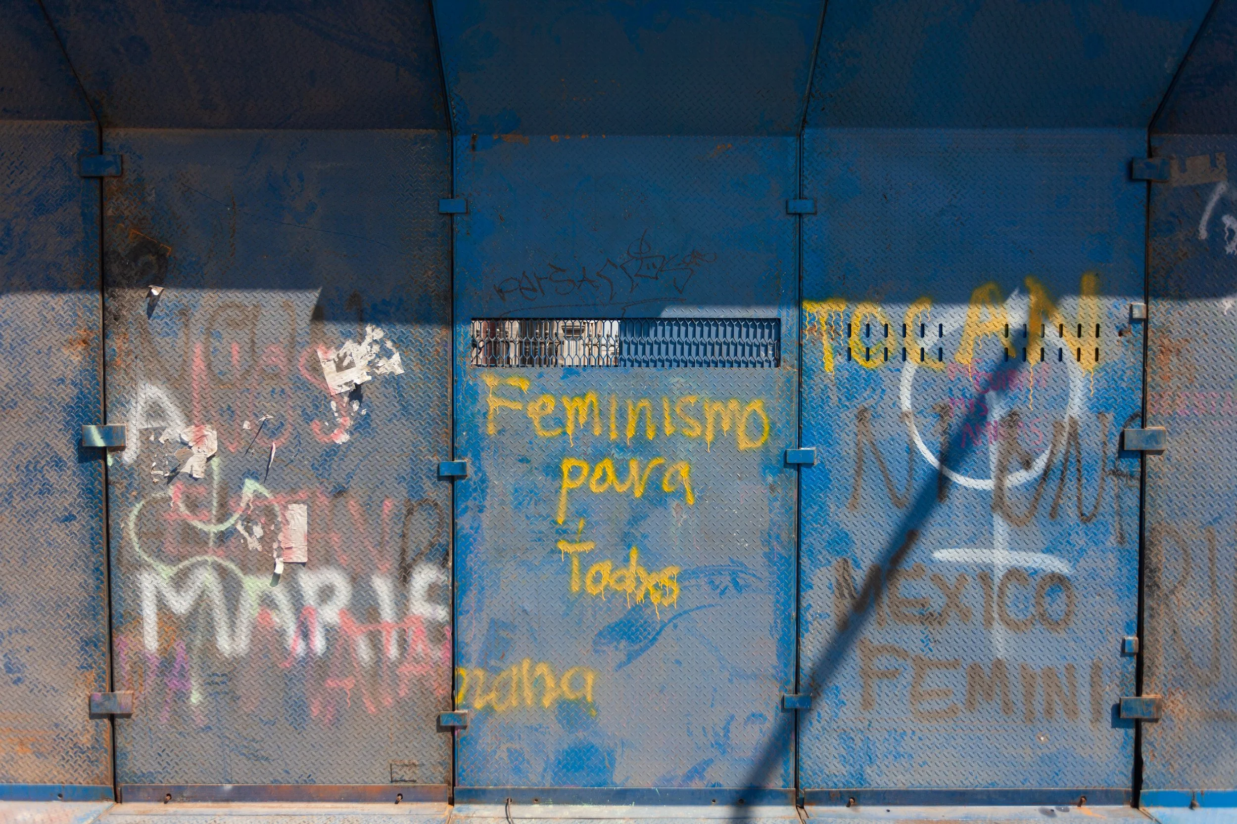 Blue metal fence with graffiti and yellow spray-painted Spanish text 'Feminismo para todas'.