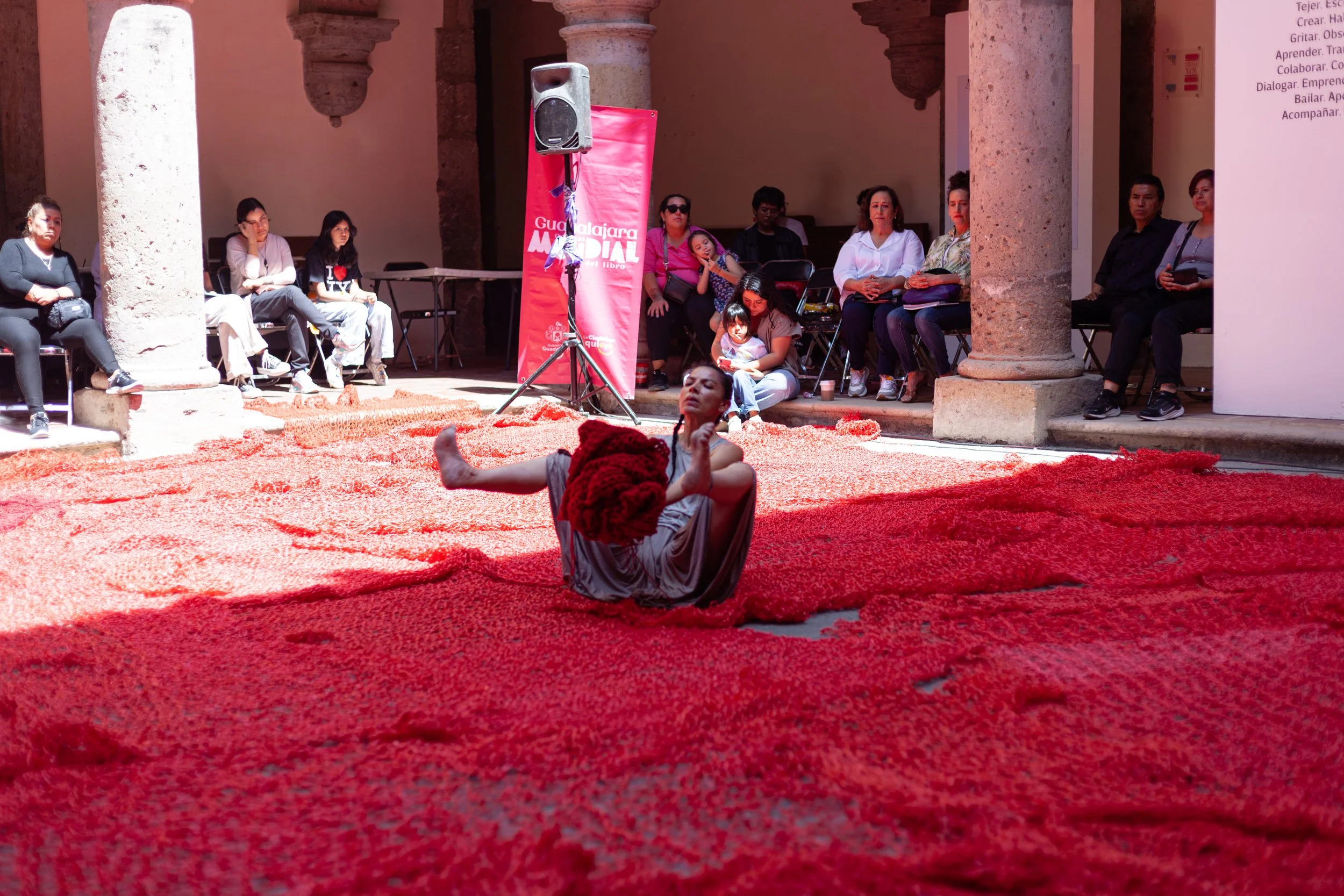 A dancer in earthy clothing performing on a large red woven textile rug in a historical building with archways, while an audience watches from seating arranged around the performance space.