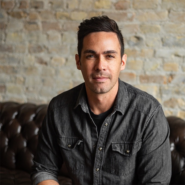 A man with dark hair and a light beard sitting on a leather couch with a brick wall background