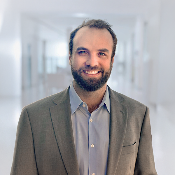 Smiling man in a gray suit and blue shirt standing in a bright, modern office space.