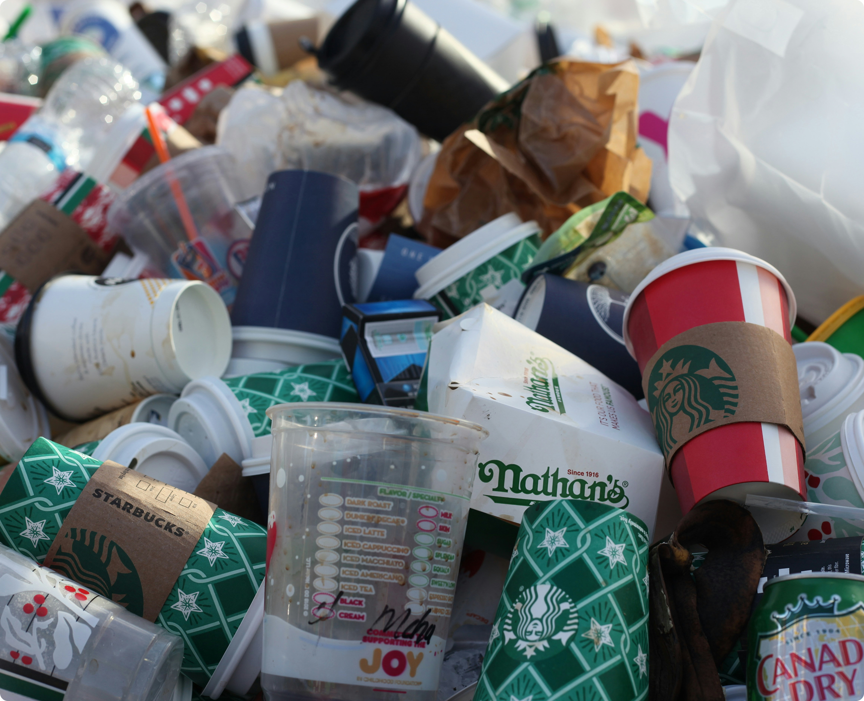 A pile of discarded paper cups, food wrappers, and plastic containers, including Starbucks and Nathan's brand, among other trash on the ground.