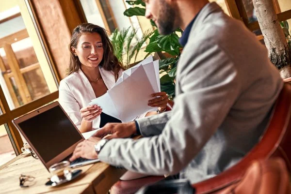 A woman and a man are in a meeting or interview in a bright office. The woman is smiling and holding documents, while the man is typing on a laptop.