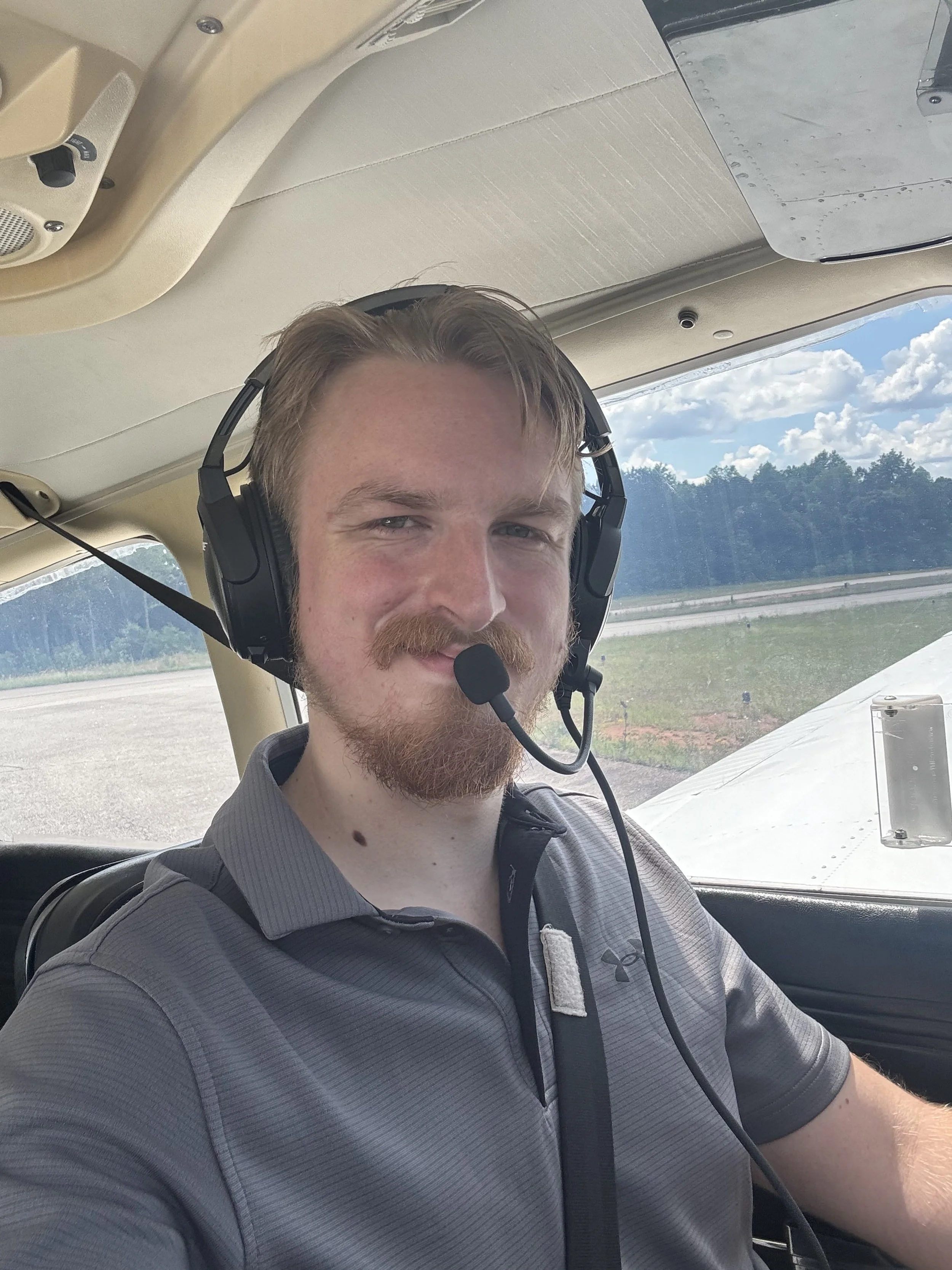 Man with blonde hair and beard wearing a gray polo shirt and headset sitting in the cockpit of a small airplane with blue sky and clouds outside the window.
