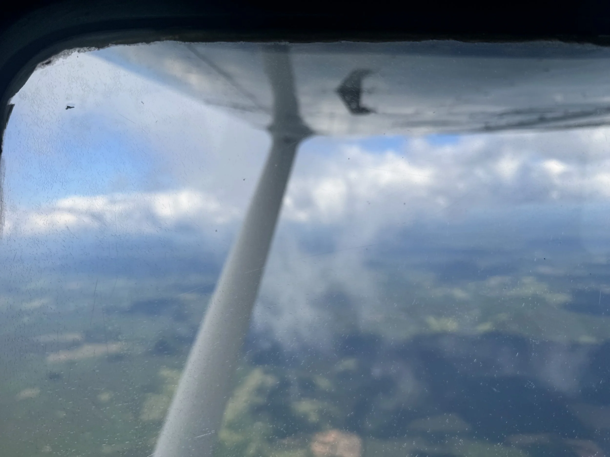 Photograph taken from inside an airplane, looking through the window at the sky with clouds and distant landscape below, showing part of the airplane's wing and strut.