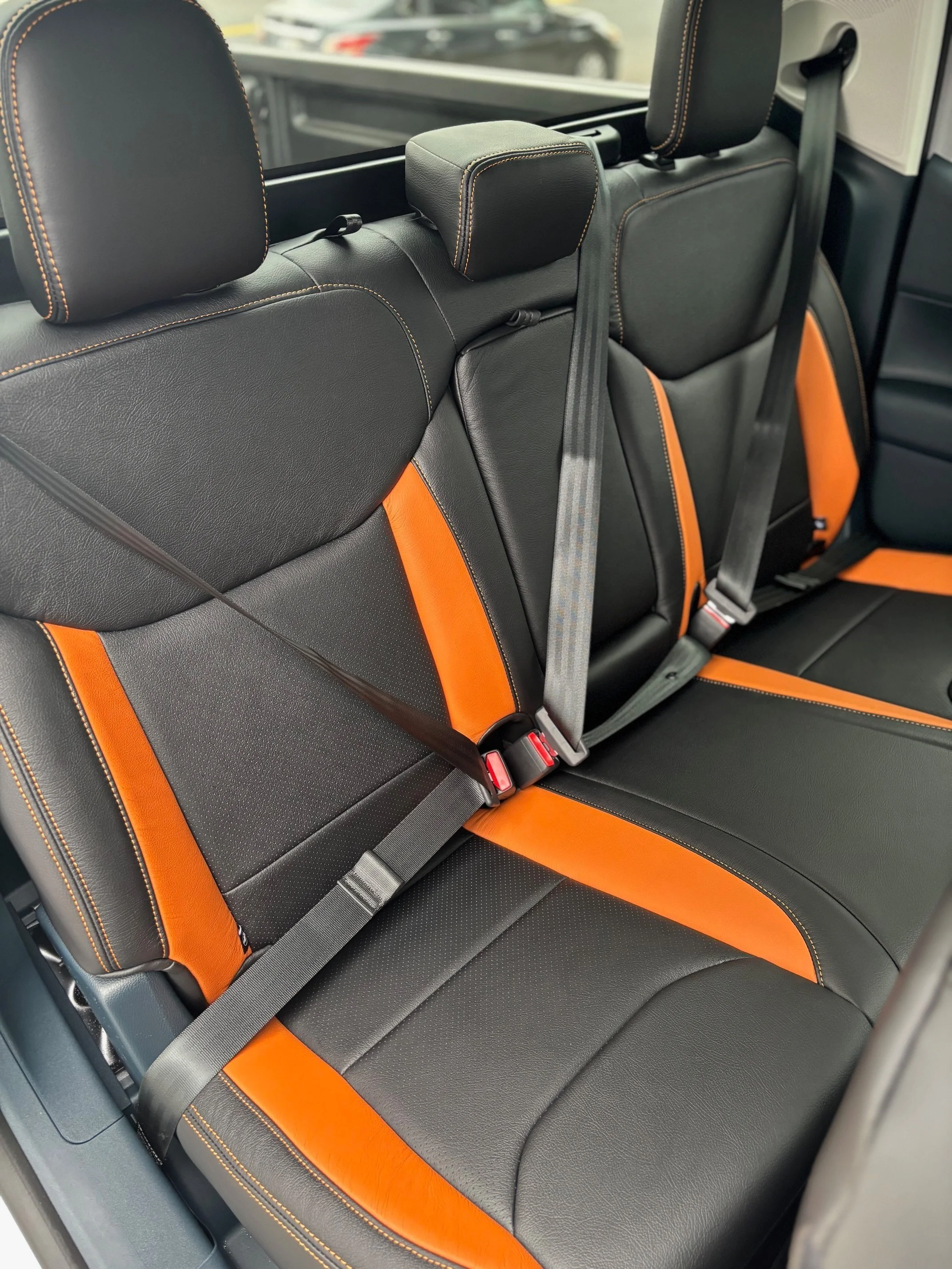 The interior of a car's back seat showing black leather upholstery with orange accents and three seat belts.