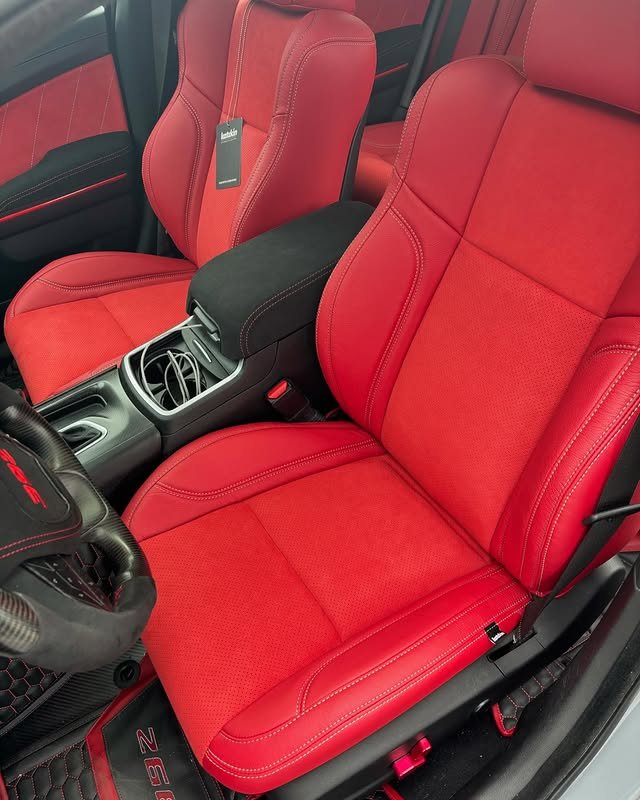 Close-up of the front seats and center console in a red sports car, showing red leather seats with detailed stitching and a black center armrest with a small compartment.