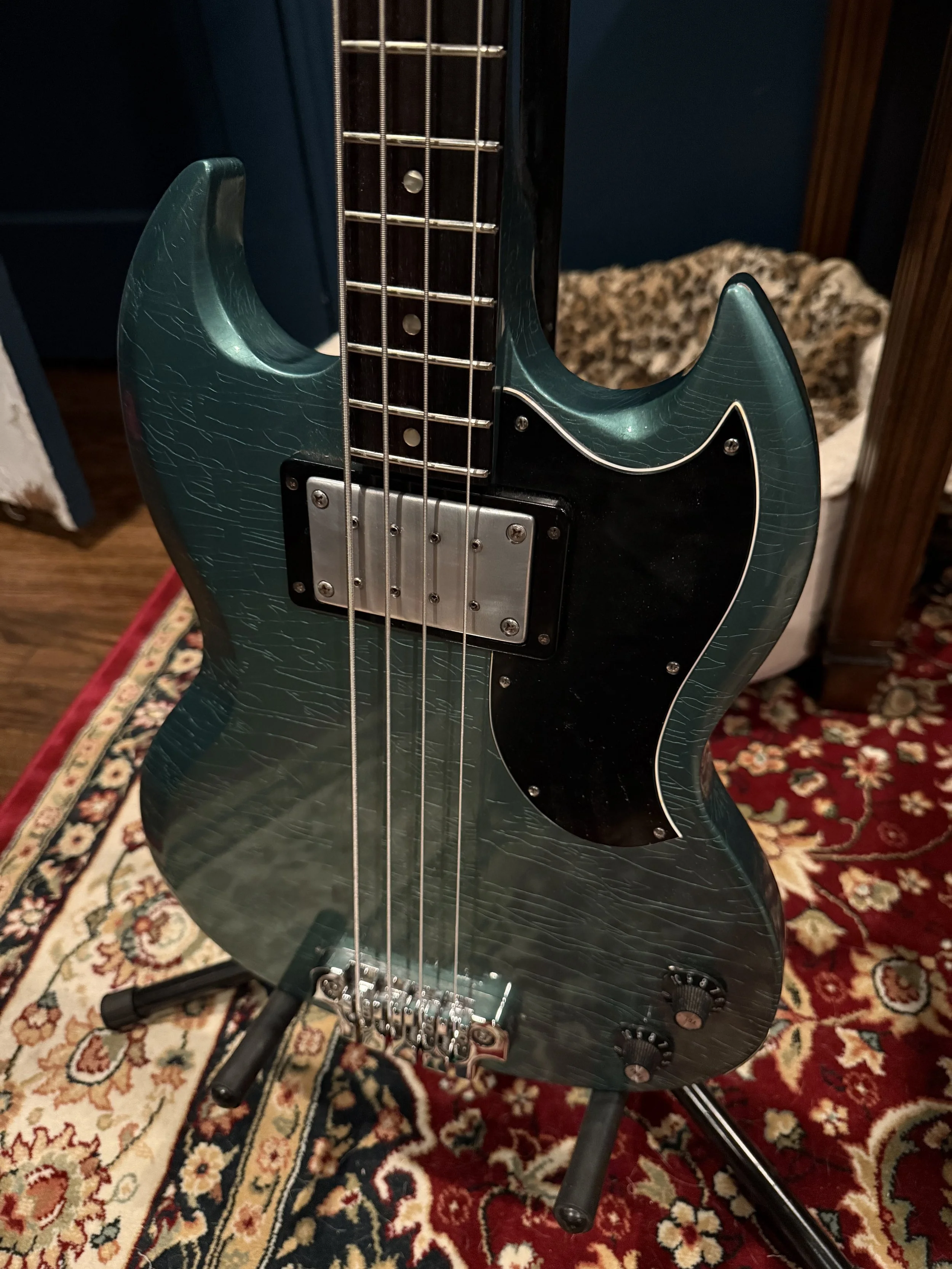 Electric bass guitar with a dark green body and a black pickguard, sitting on a stand, with a patterned rug underneath.