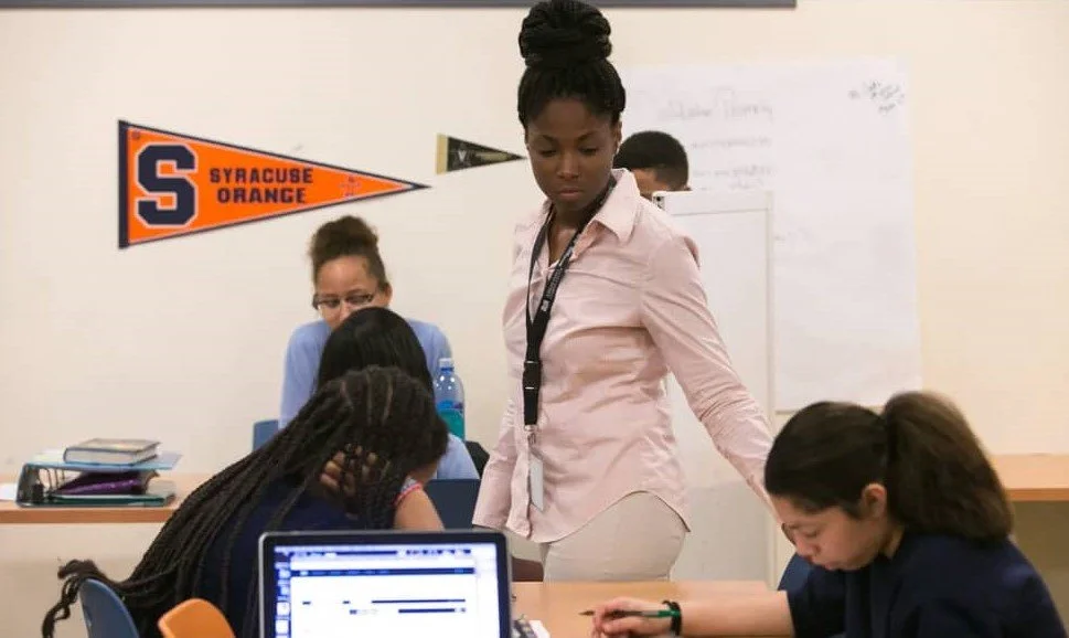 Teacher supervising students in a classroom setting with a Syracuse University pennant, promoting academic mentorship"
