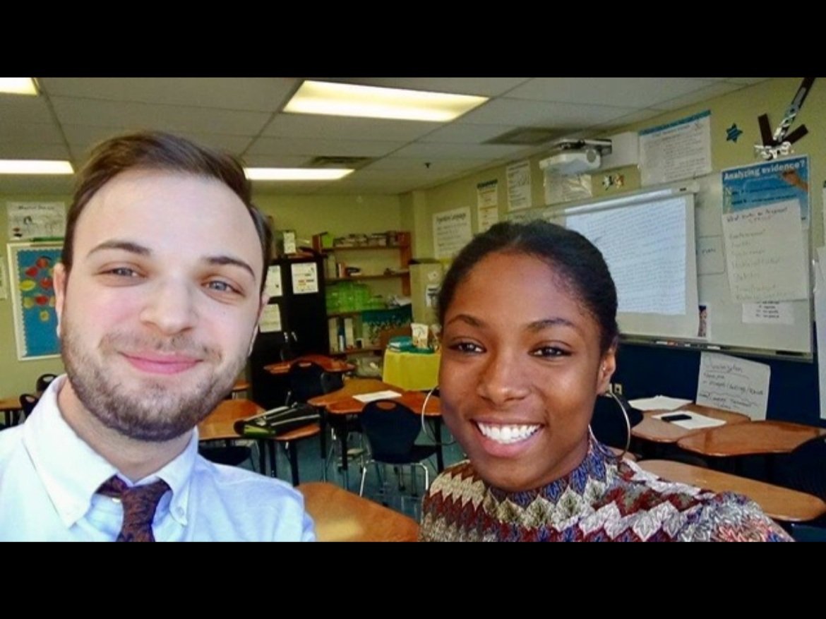 A young man and woman smiling for a selfie in a classroom with desks, whiteboard, and educational posters in the background.