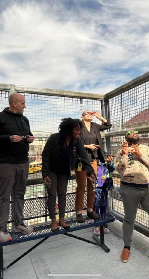 Four people standing on a rooftop with a metal fence; three are on a bench, one is taking a photo, layered clouds in the sky.