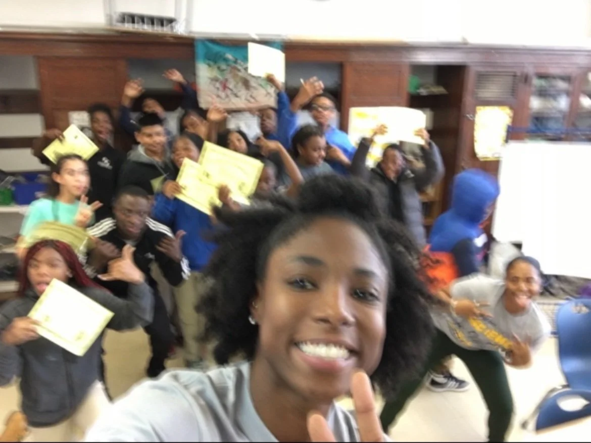 Smiling African American teacher in front of blackboard with children at desks and a banner reading 'Exceptional Teacher Resident: Ms. Evans