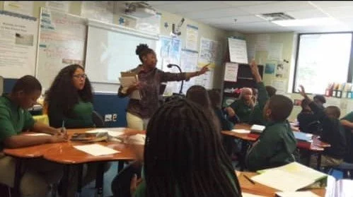 A teacher leading a classroom discussion with students raising their hands.