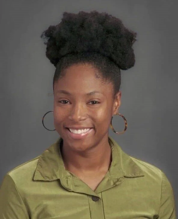 Portrait of a woman with natural hair styled in a large puff, wearing hoop earrings and an olive-green shirt, smiling against a gray background.