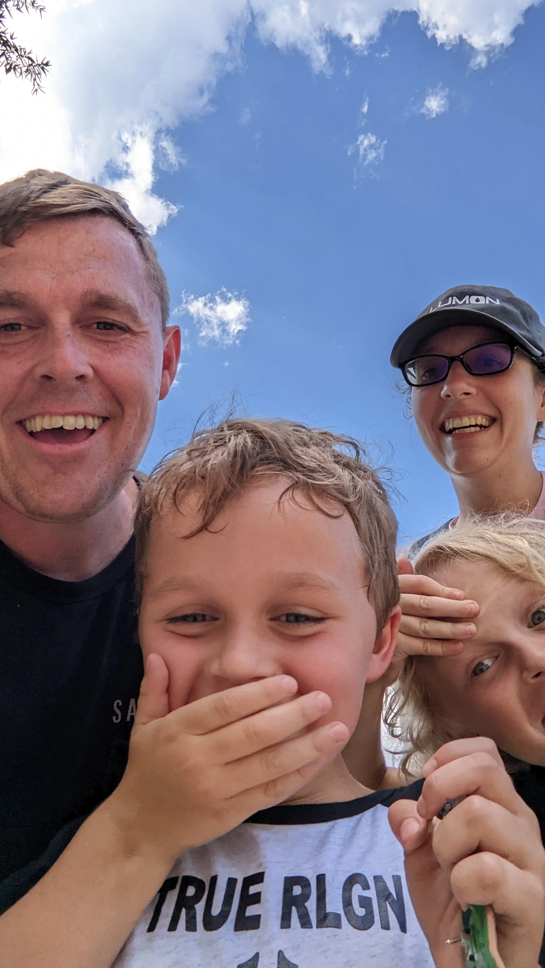 A group of five people, including two adults and three children, smiling and making playful poses against a bright blue sky with some clouds.