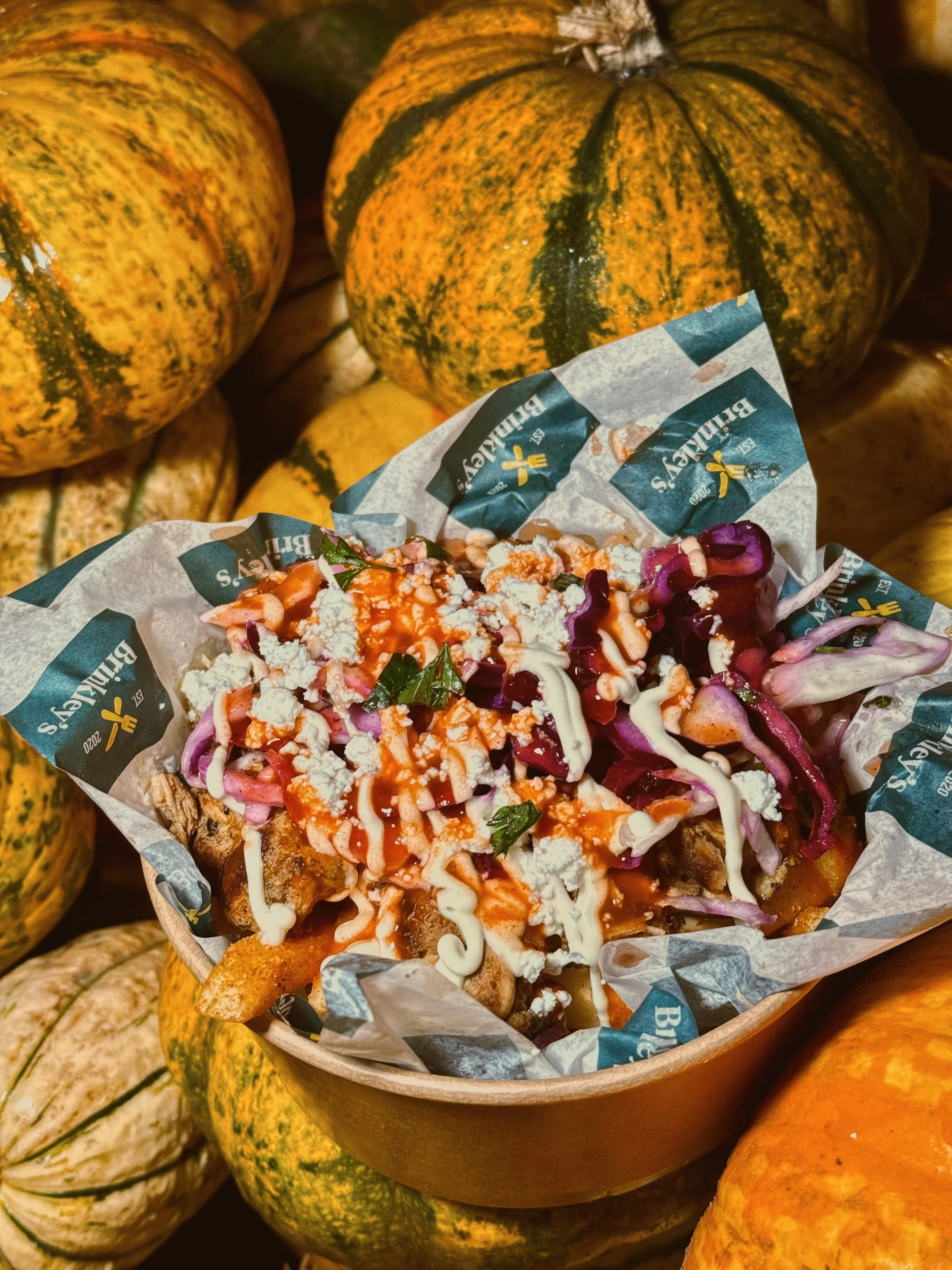 A bowl of loaded fries topped with shredded cheese, purple cabbage, green herbs, and drizzled with white and red sauces, surrounded by small orange and green striped pumpkins.