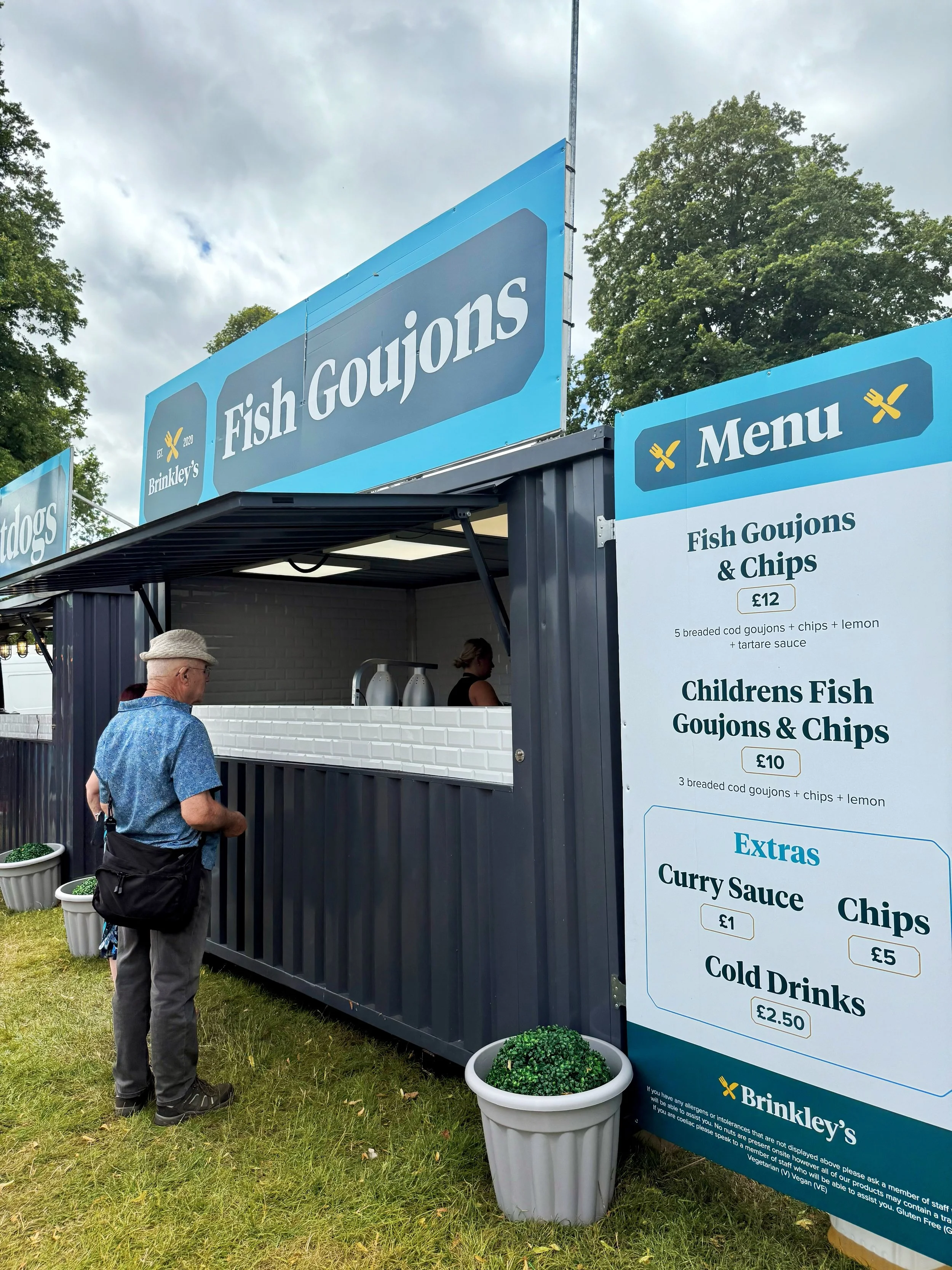 Brinkley's Fish Goujons street food trailer serving freshly cooked breaded cod goujons and chips at an outdoor event, with a customer ordering from the blue-branded catering unit beside a clearly displayed menu.