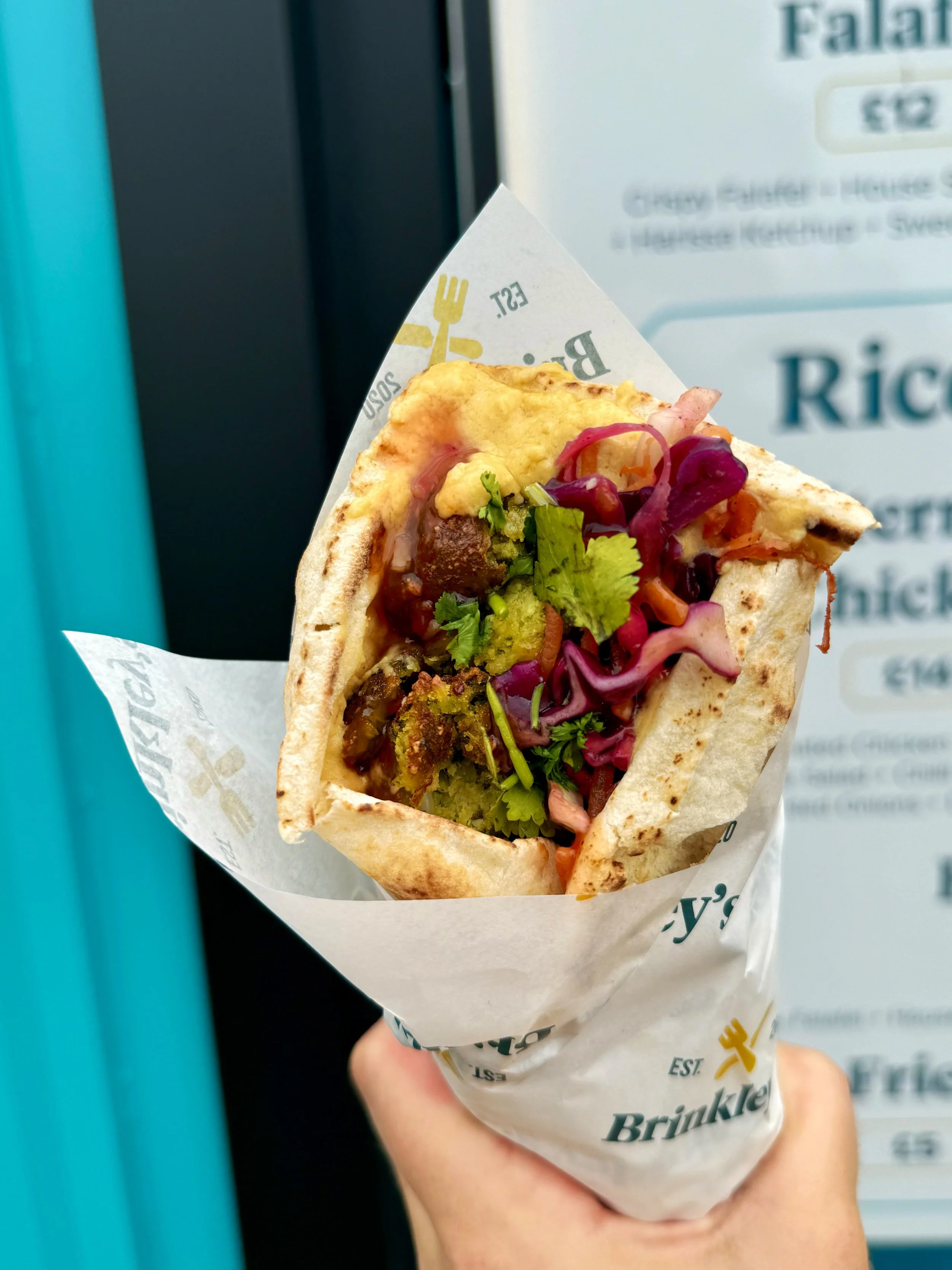 Hand holding a taco with colorful vegetables, grilled meat, and sauce, wrapped in branded paper from Brinkley's.