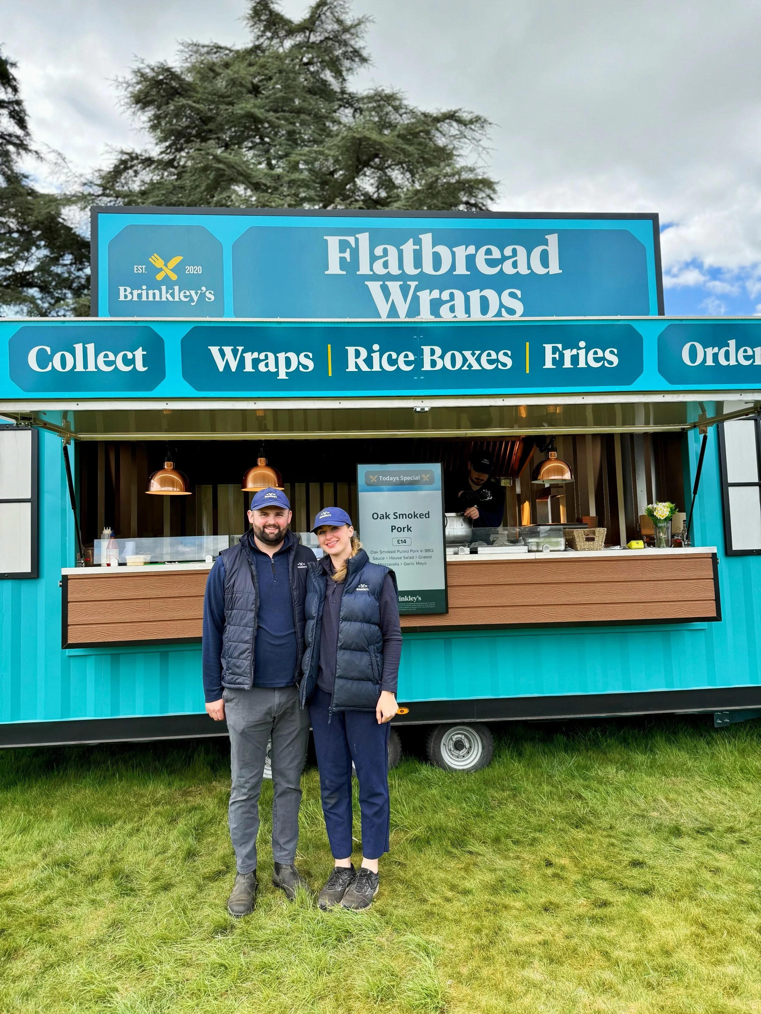 Two people stand in front of a teal and brown food truck labeled 'Flatbread Wraps' with a menu including wraps, rice boxes, and fries. They are smiling and wearing navy blue jackets and caps, with a grassy area and trees and a cloudy sky in the backg