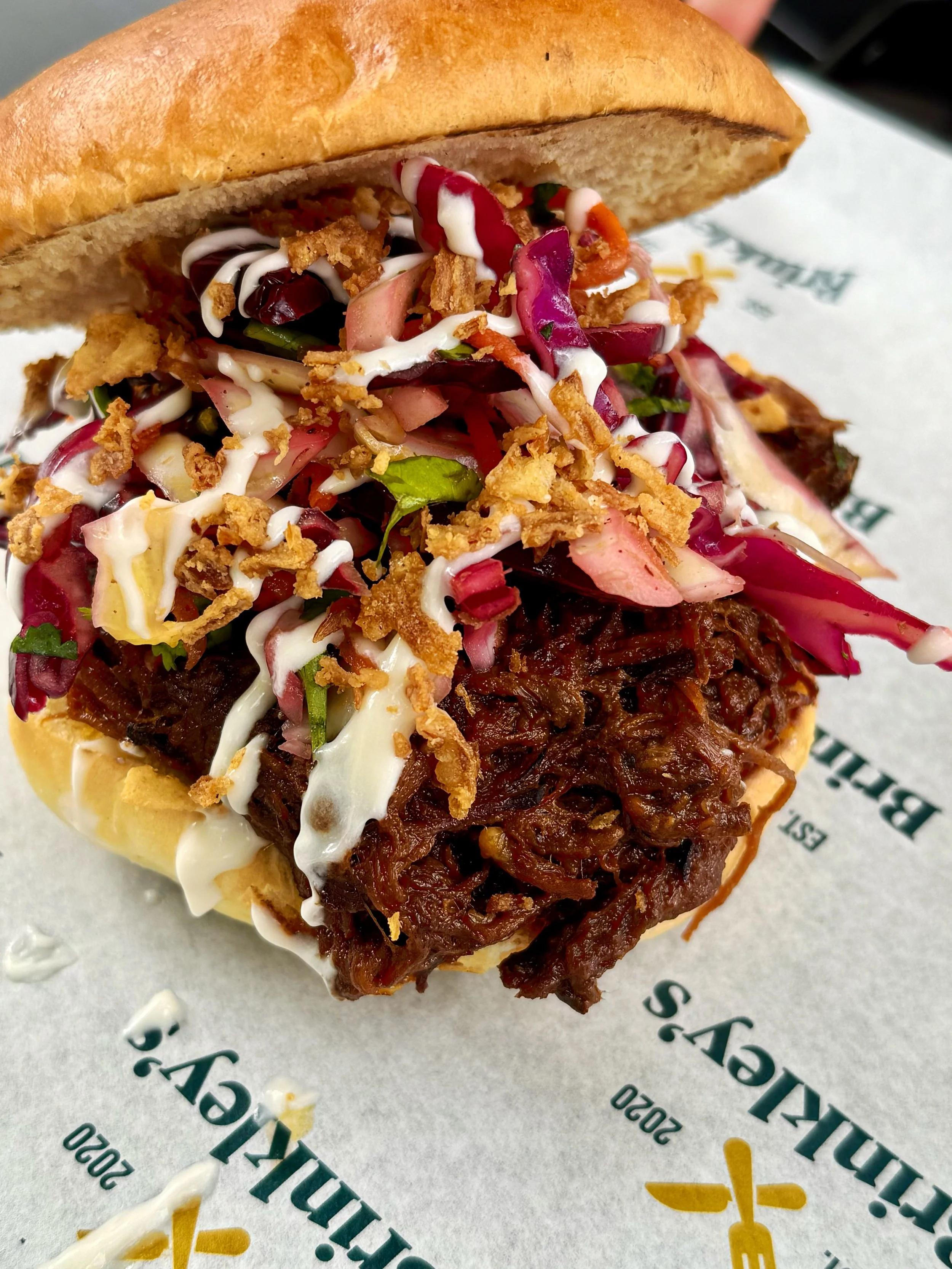 Close-up of a barbecue sandwich with shredded meat, coleslaw, and sauce in a sandwich bun, on branded paper.