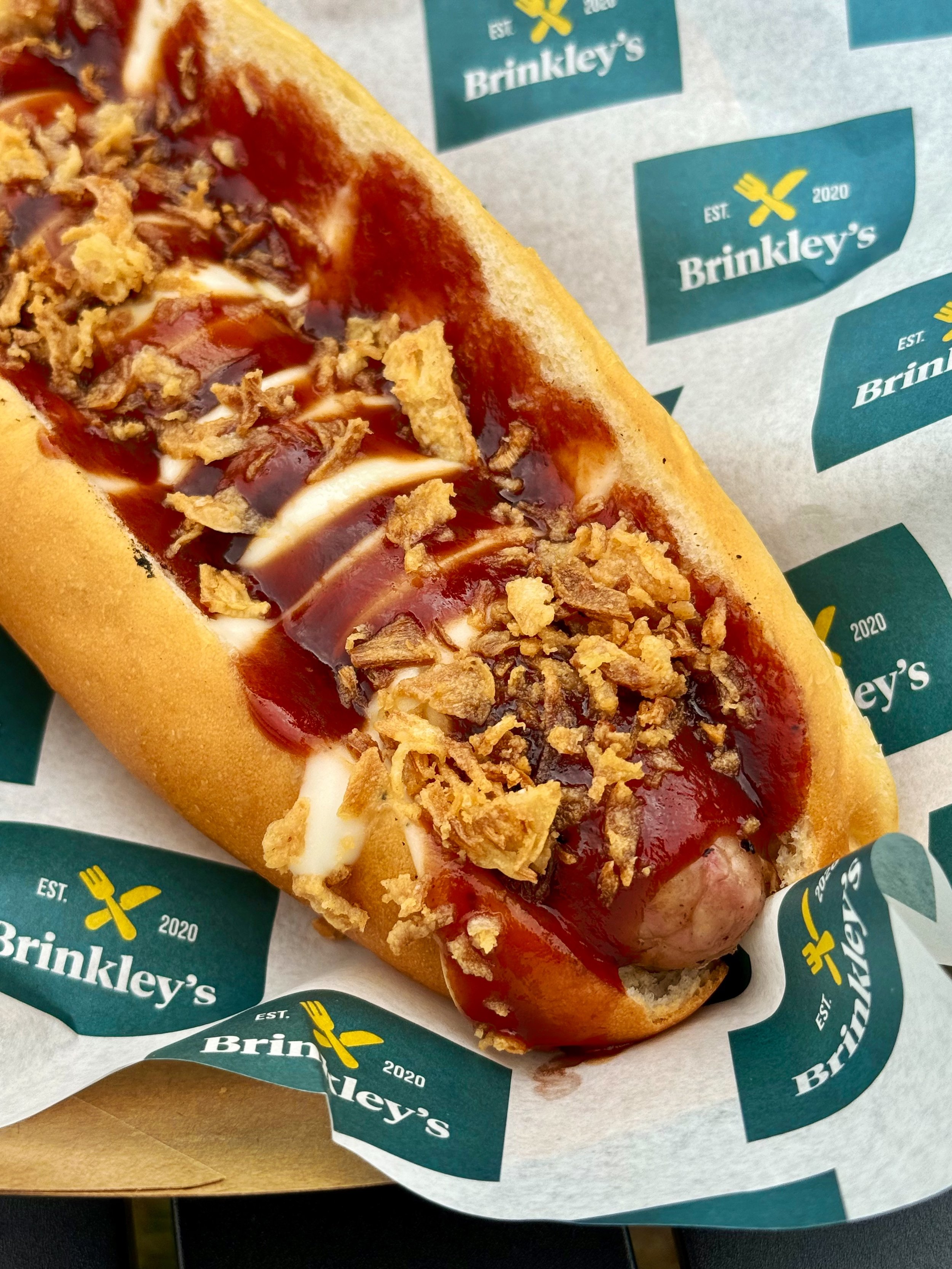 Hot dog topped with homemade chilli, drizzled in sour cream, and crispy fried onions in a bun on branded paper.