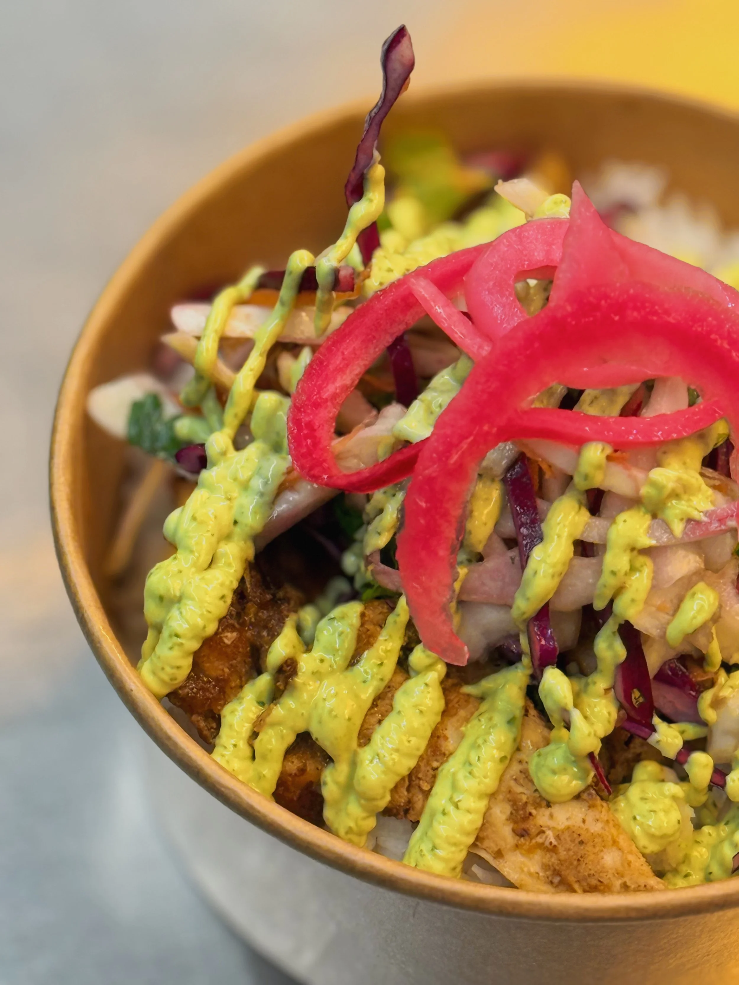 A bowl of crispy fried fish topped with sliced red peppers, shredded cabbage, and drizzled with green avocado sauce.