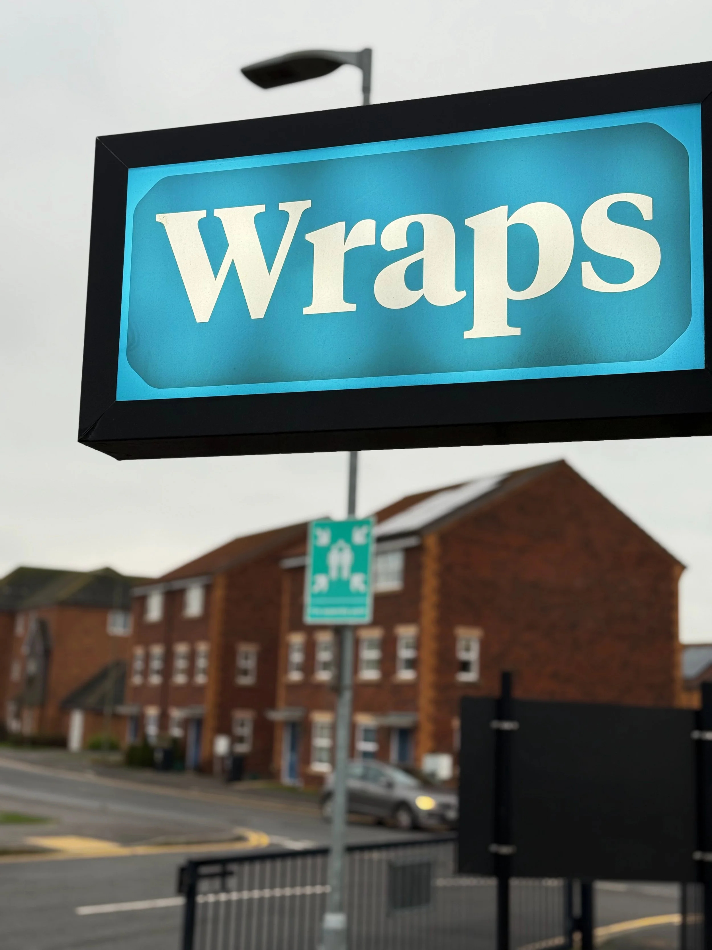 Close-up of a lit sign with the word 'Wraps' in white text on a blue background, hanging outside near residential buildings.