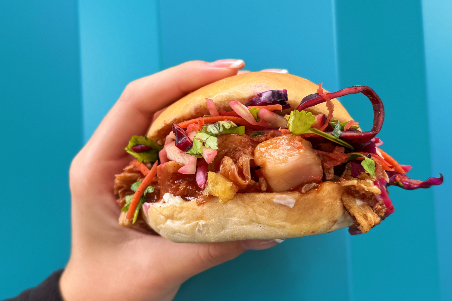 Hand holding a sandwich with pulled pork, coleslaw, and mixed vegetables in a bun against a teal background.