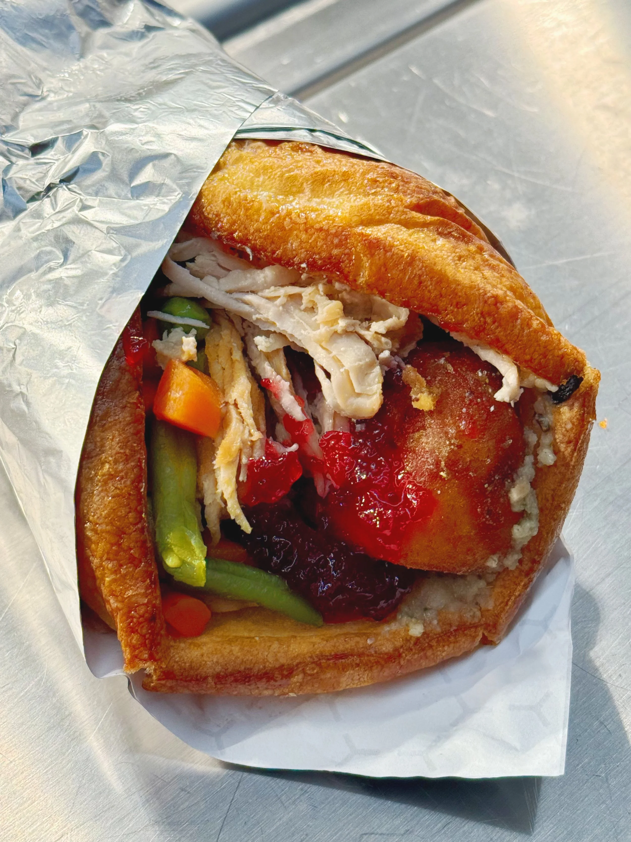 A partially unwrapped turkey and stuffing sandwich with vegetables and cranberry sauce, served on a bun.