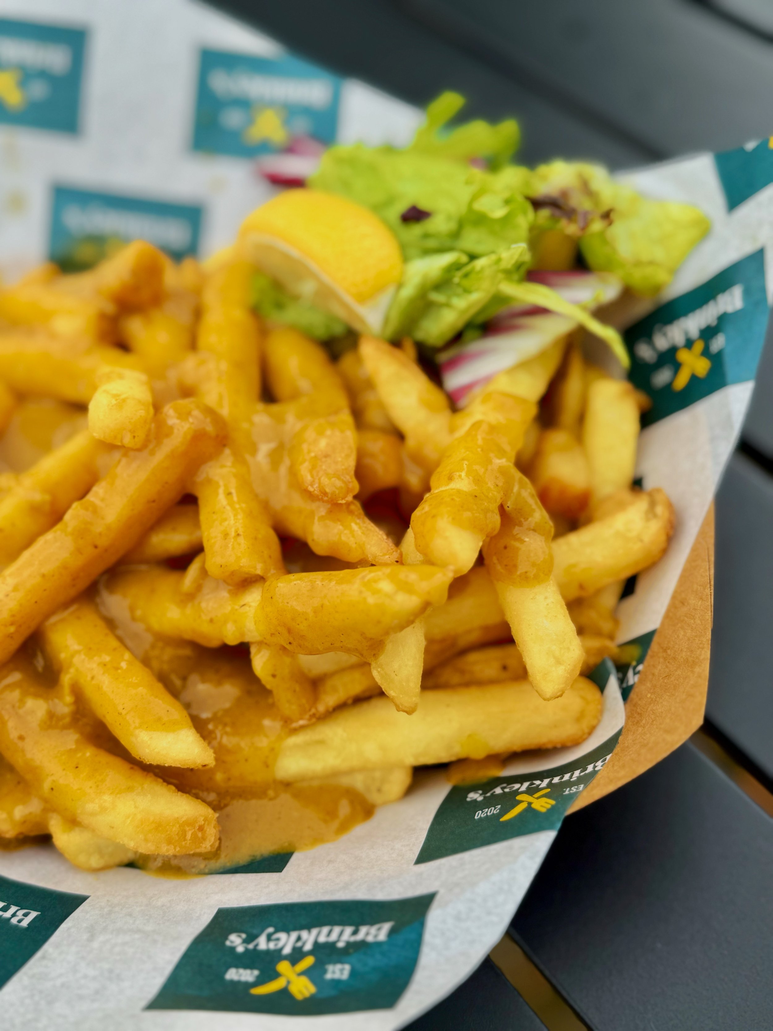 French fries with curry sauce, with a small salad of lettuce and a lemon wedge, in a paper container with Brinkley's logo.