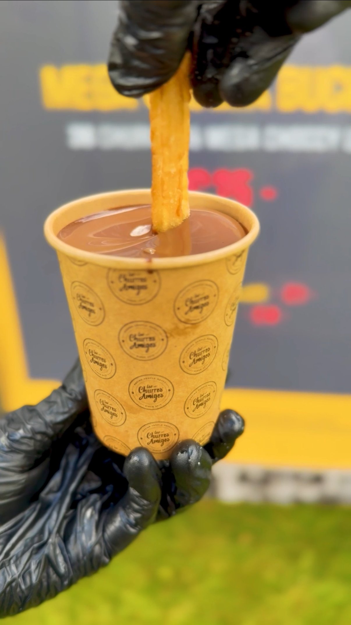 A hand wearing a black glove holding a paper cup filled with melted chocolate and a churro dipped in the chocolate.