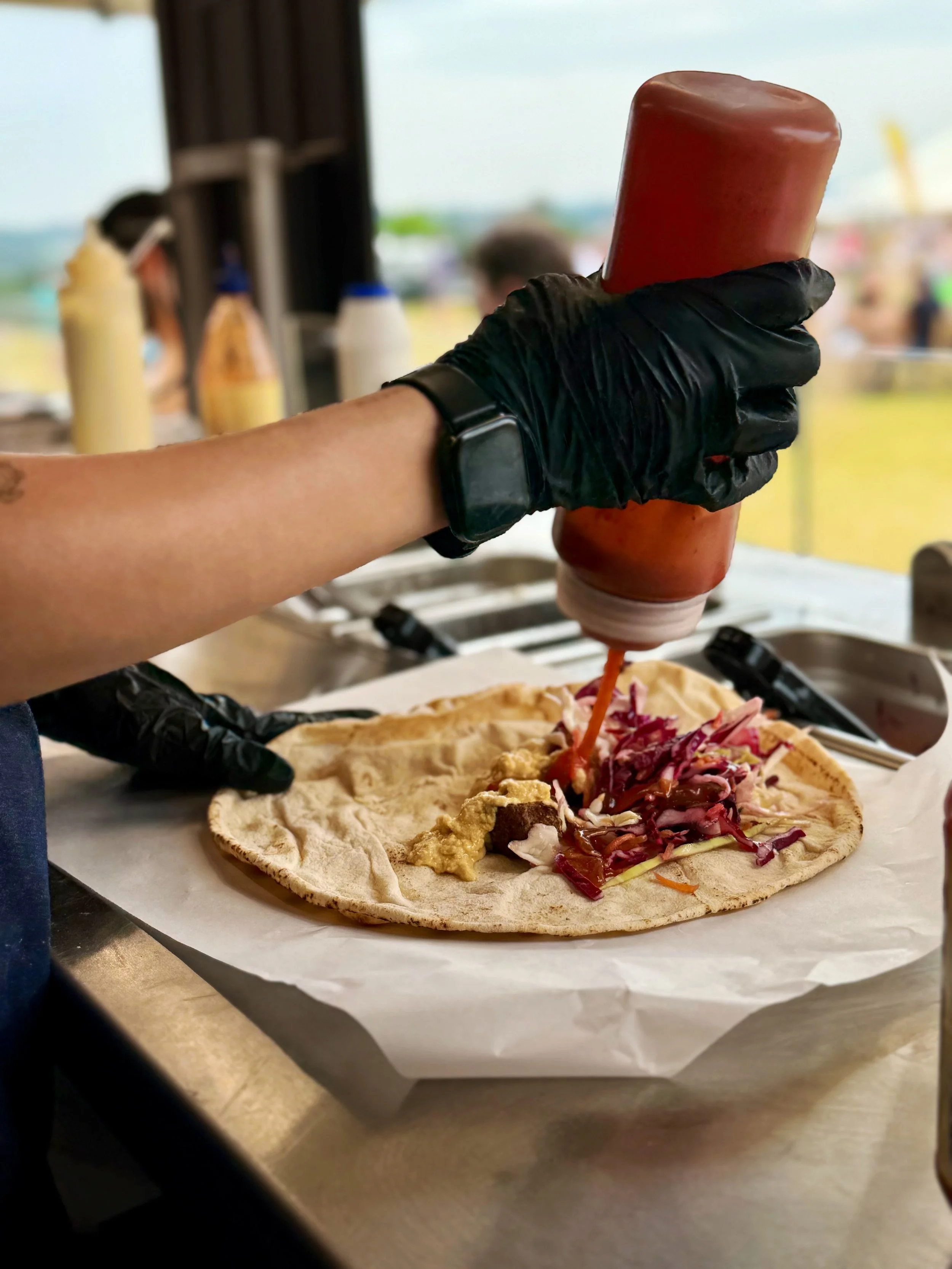 Person wearing black gloves adding ketchup to a hot dog on a flatbread wrap at a food stand.