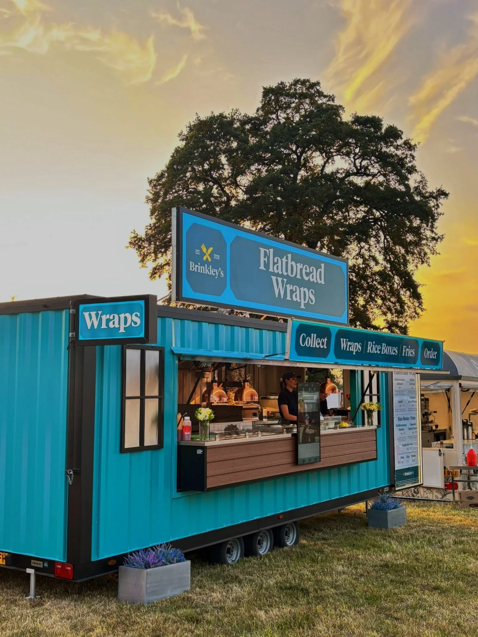 A food stand named 'Brinkley's' serving flatbread wraps at sunset, with a large tree and a yellow-orange sky in the background.