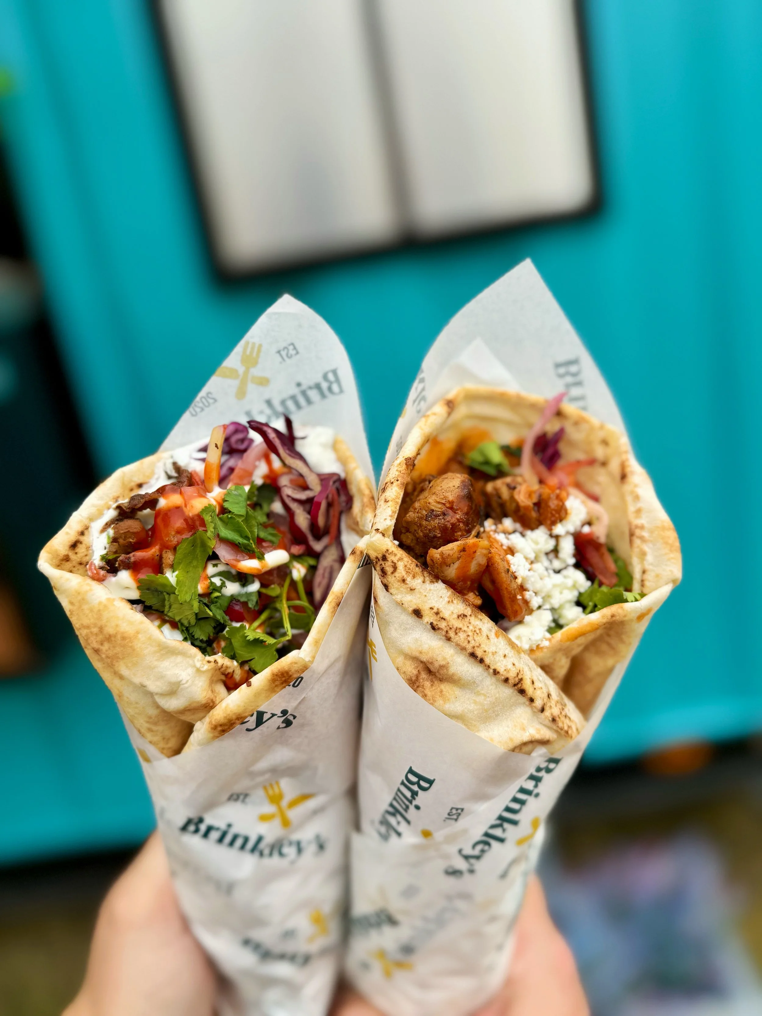 Two beef shawarma wraps in branded paper, filled with meat, vegetables, and sauces, held up against a blue background.
