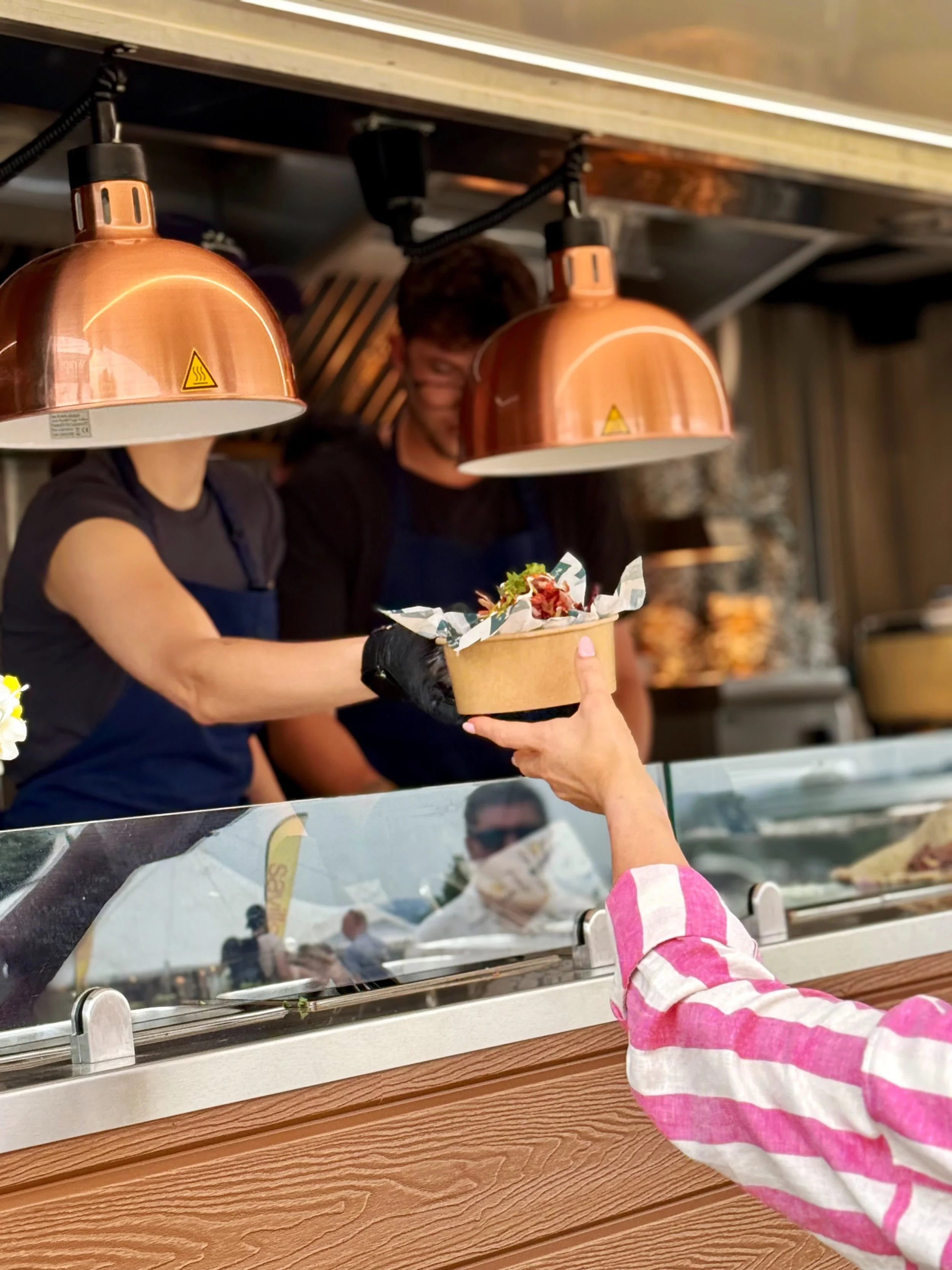 Person in pink and white striped shirt receiving a food order from a worker in a kitchen with copper pendant lights.