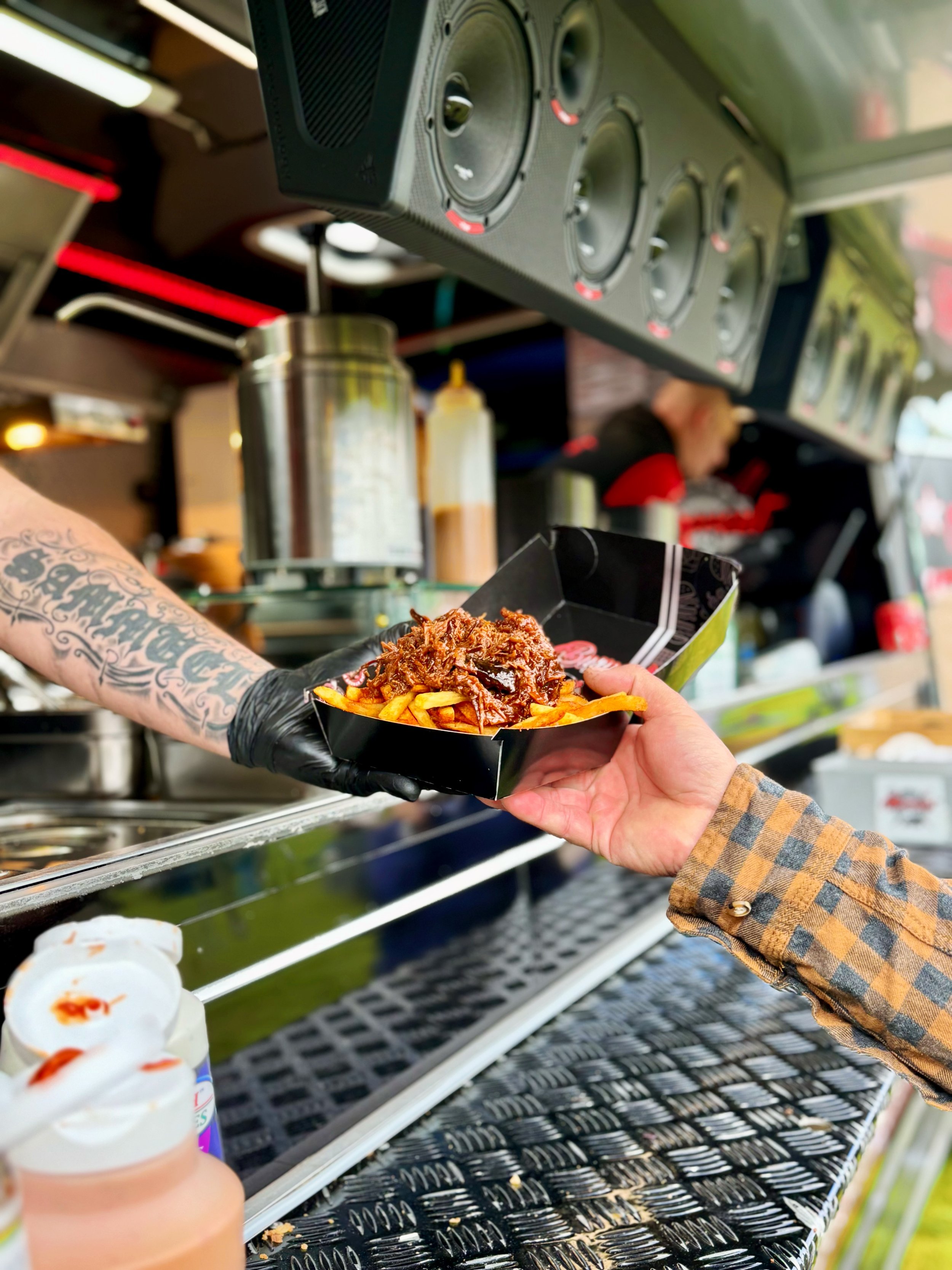 A person in a plaid shirt is receiving a serving of French fries topped with pulled pork from a food truck. The truck has a metal counter and visible equipment in the background.