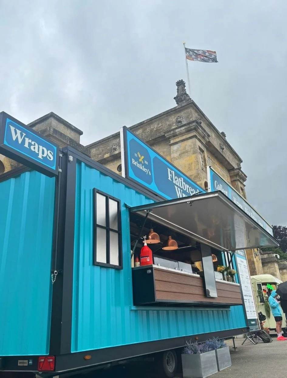Blue food truck with service window, fire extinguisher, and menu, parked in front of a historic building with a flag on top.