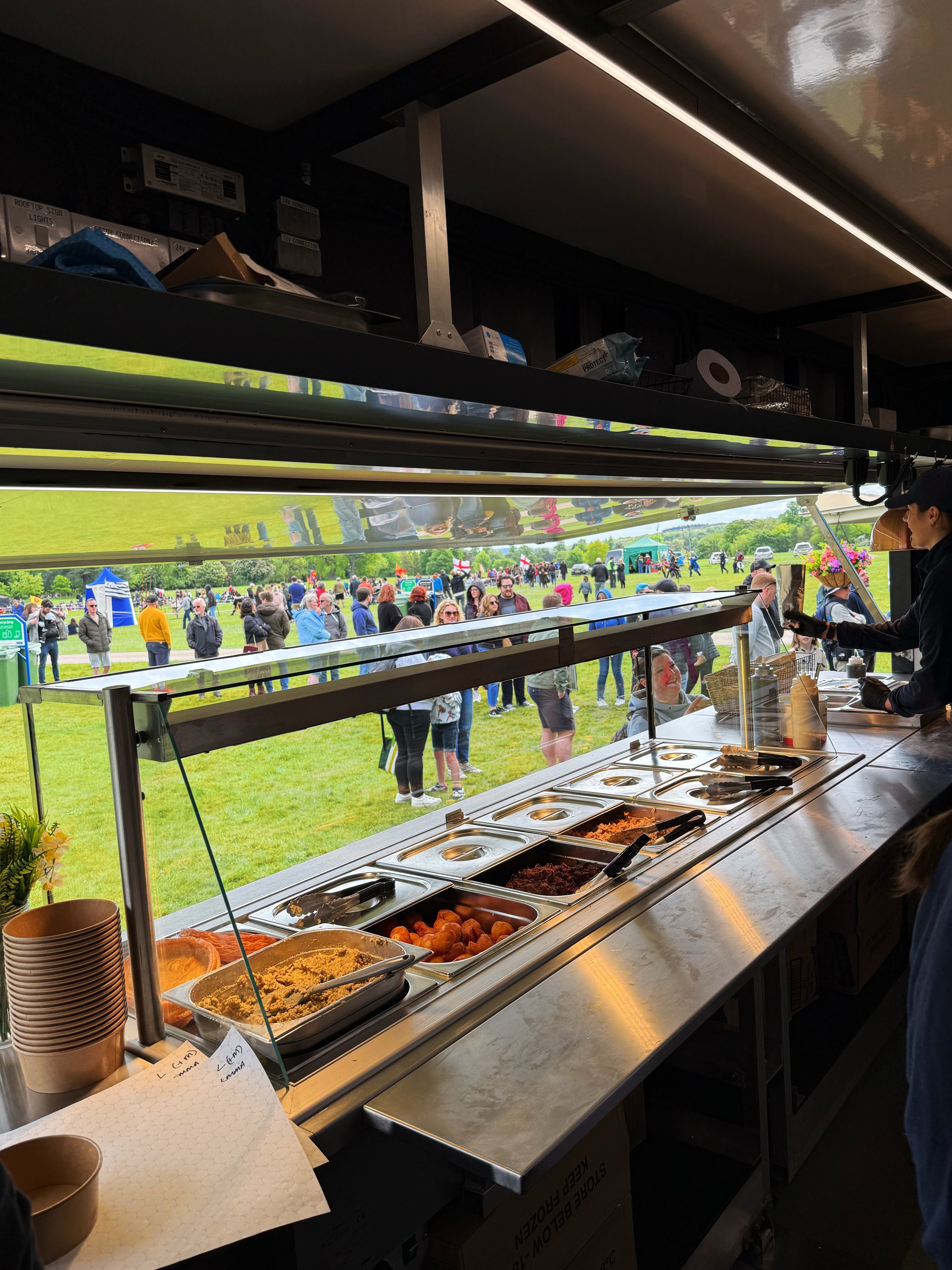 View of a food service counter at an outdoor event with a line of people waiting outside, the counter has various food trays, and the scene includes a grassy field and visitors in the background.