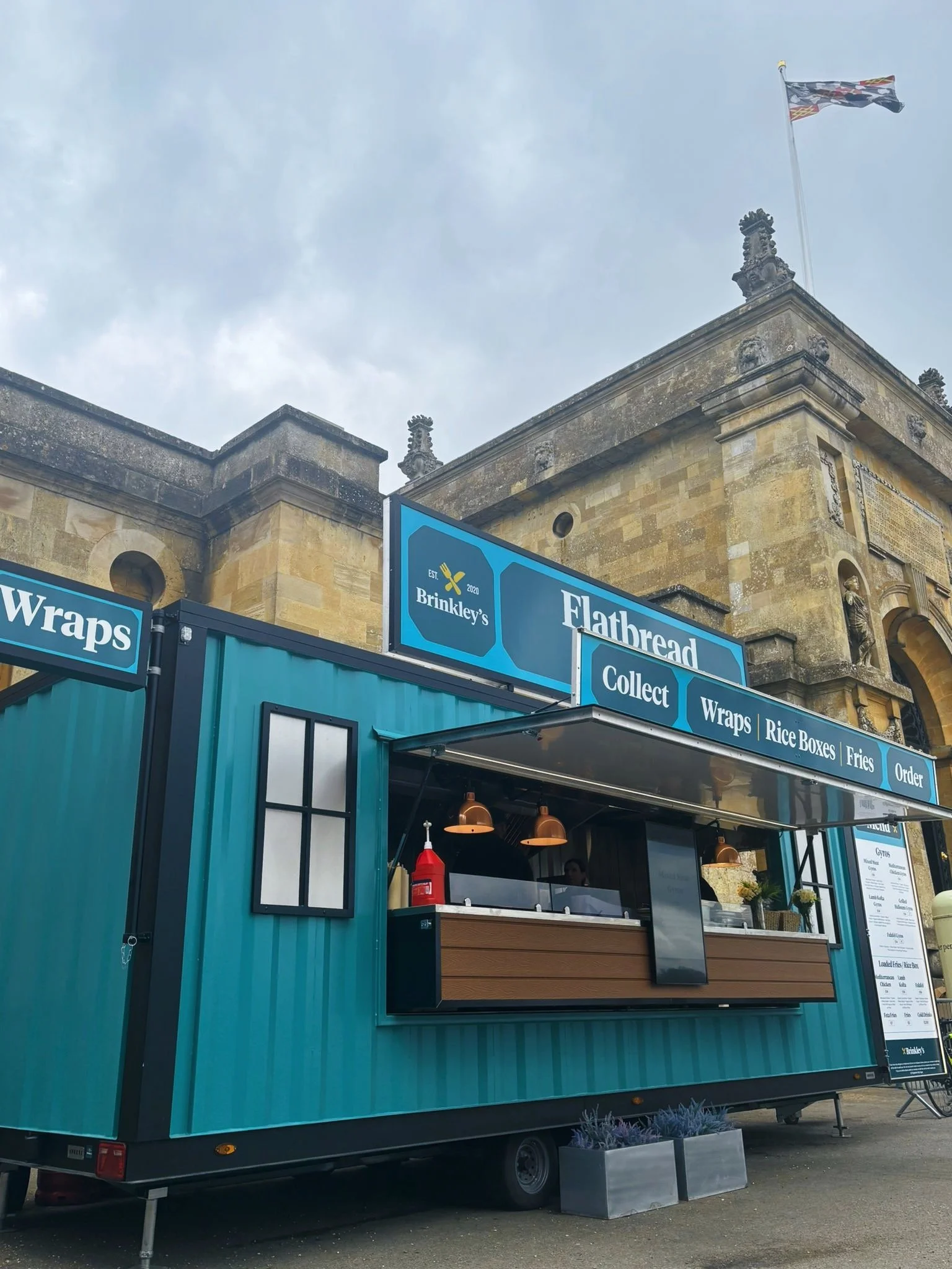 A teal food truck named 'Flatbread' is serving at an outdoor location, with a menu sign listing items like wraps, rice boxes, fries, and order options. An old stone building with decorative elements and a flag on top is visible behind the truck.
