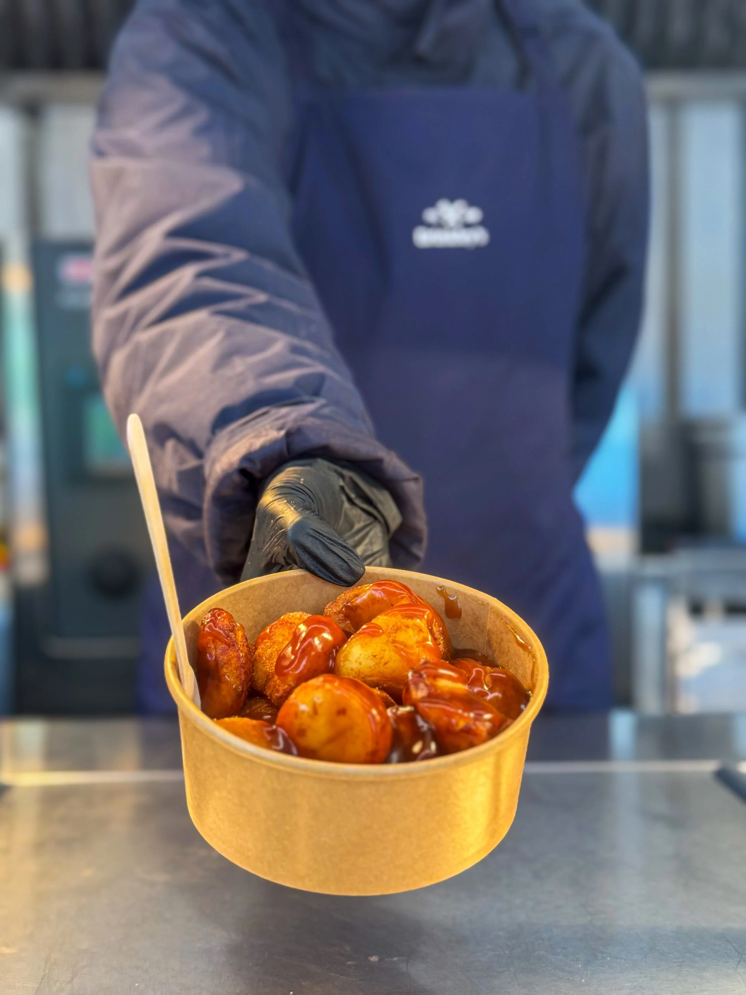 Person wearing a dark jacket and black gloves presenting a cup of glazed caramel apples with a wooden stick in a food service setting.