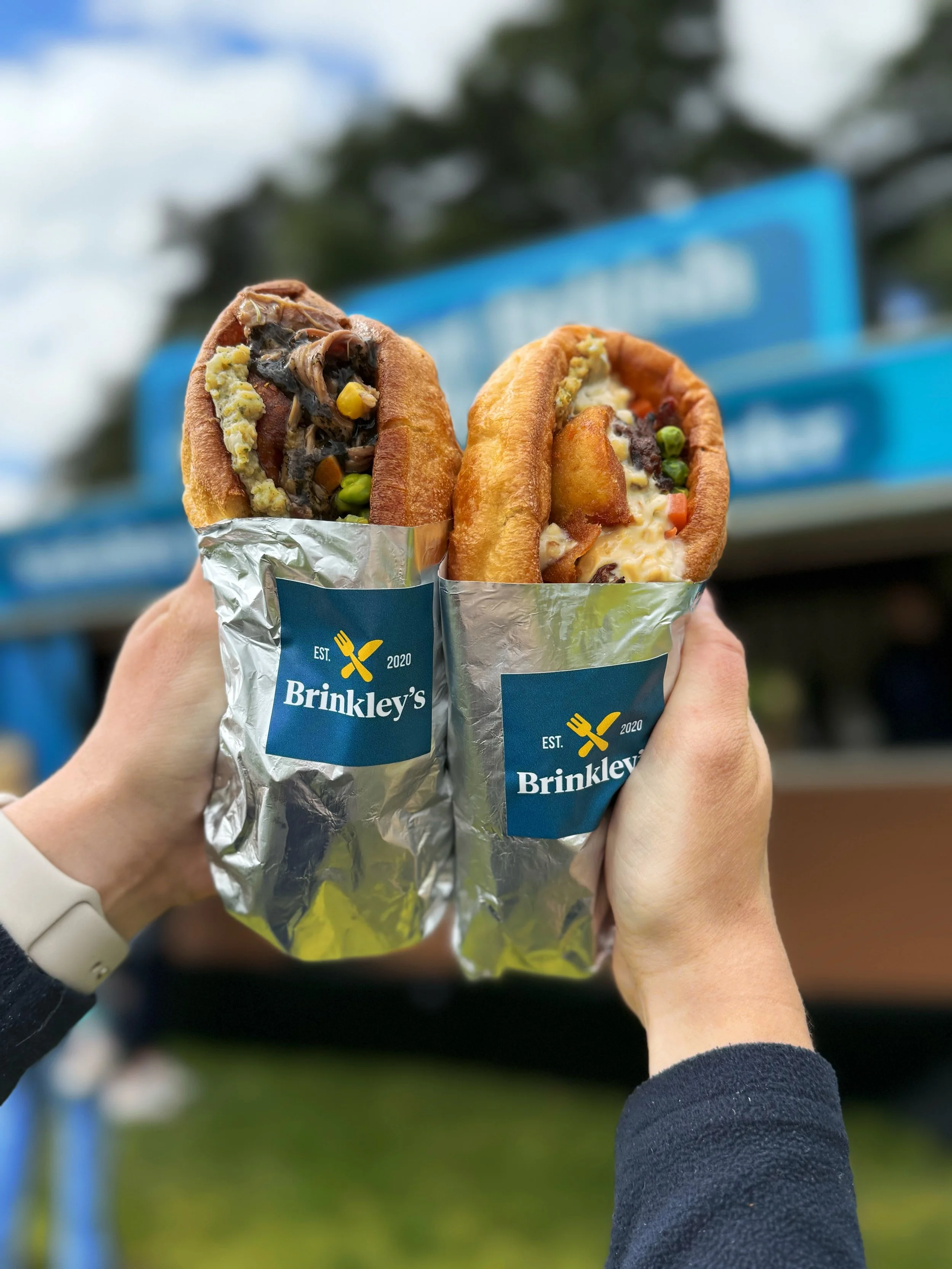 Two hands holding hot dogs wrapped in foil with a blue Brinkley's label. The hot dogs are topped with various ingredients and are outdoors in front of a blue sign and partly cloudy sky.