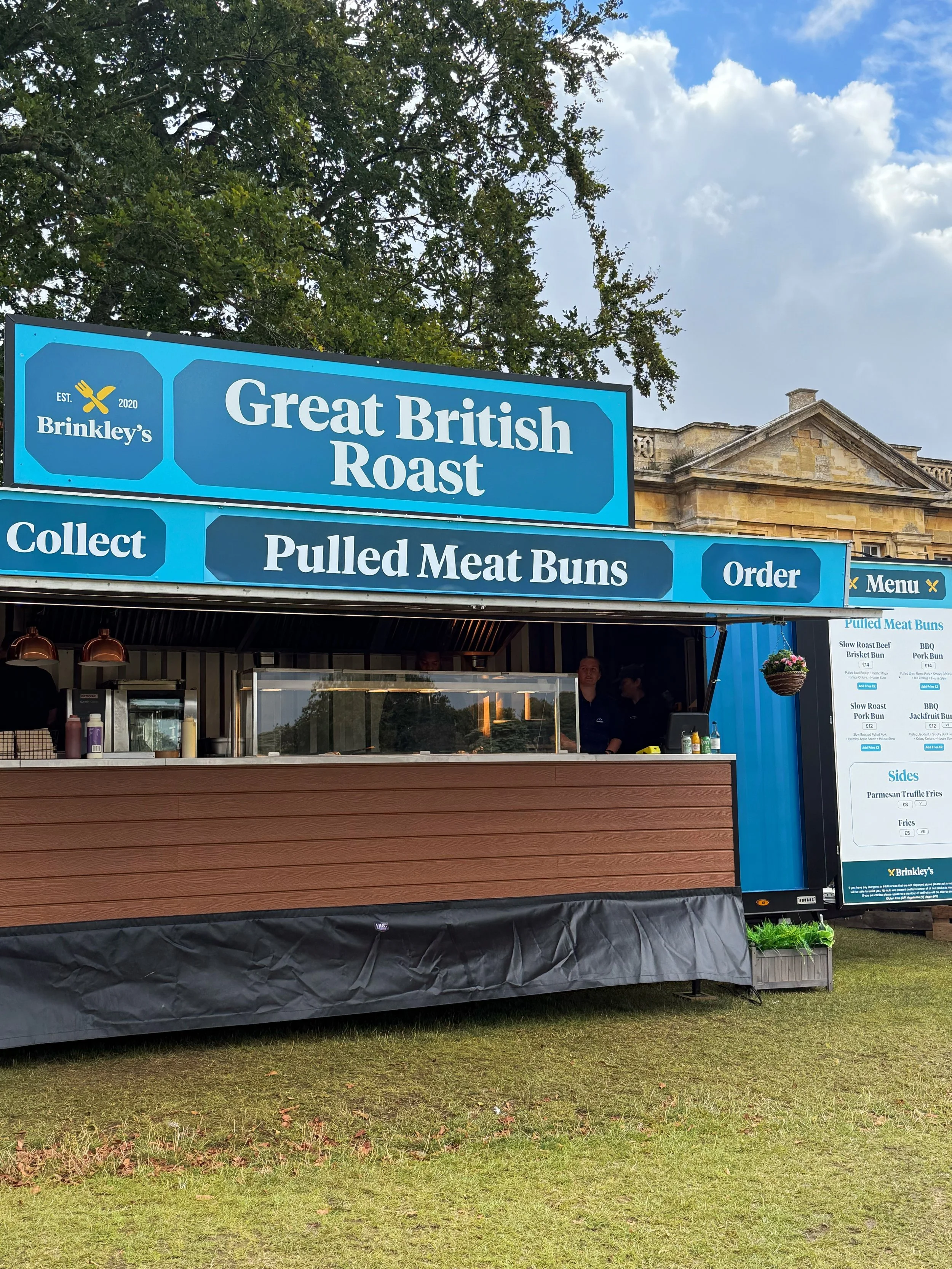 Food stand with a blue sign that reads 'Great British Roast' and 'Pulled Meat Buns'. The stand offers various roasted meats and sides, with a menu board visible on the right.